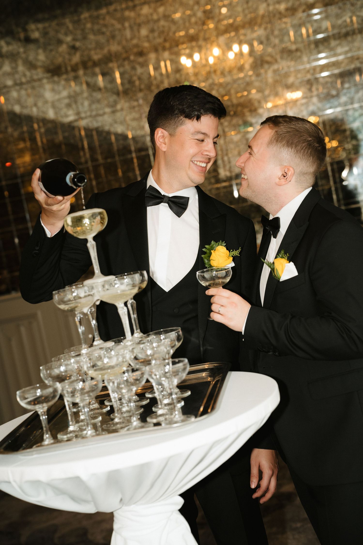 Two men in tuxedos share a laugh while stacking champagne glasses into a tower at an elegant reception.