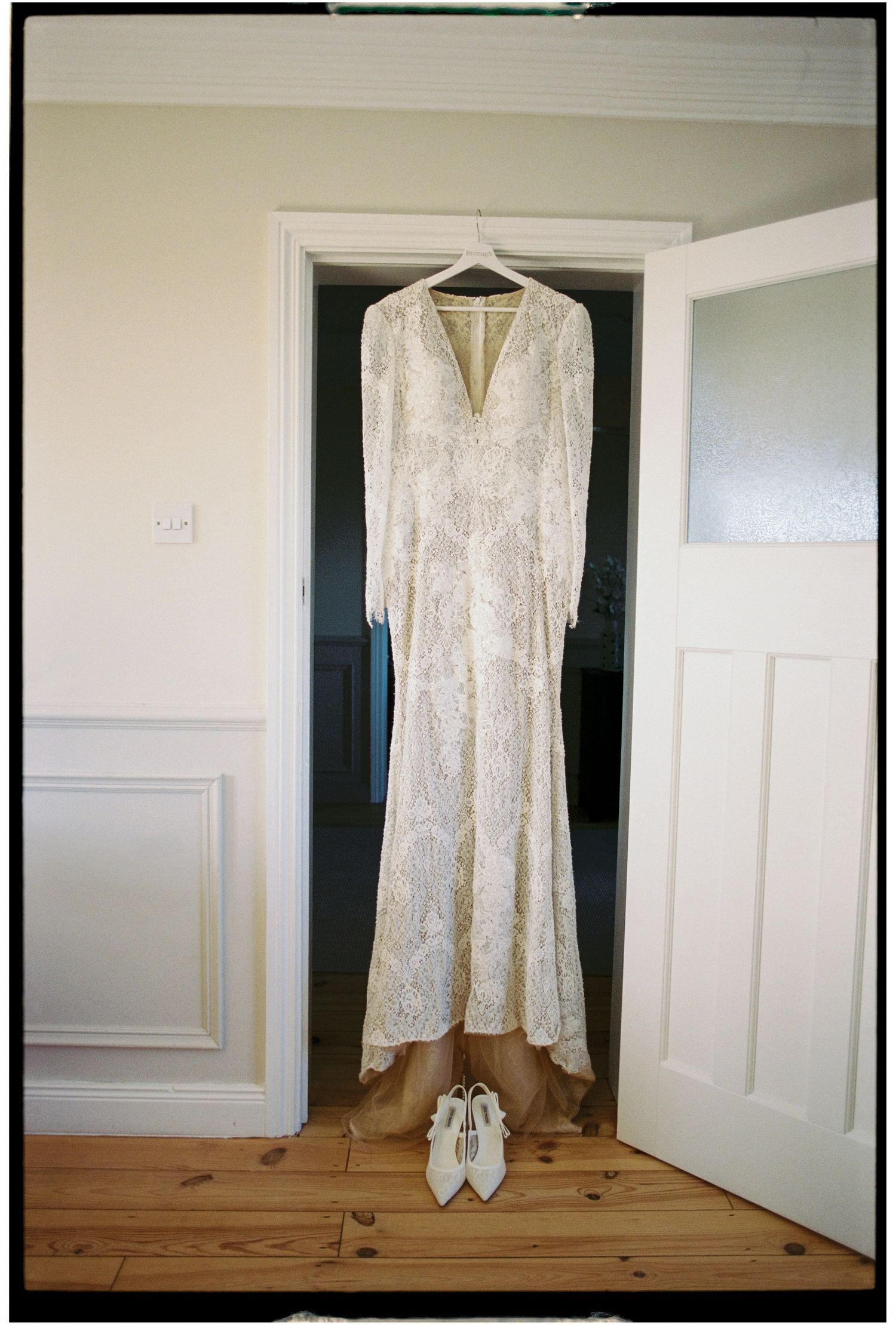 A vintage-style lace wedding dress with long sleeves hangs on a door with white shoes placed underneath.