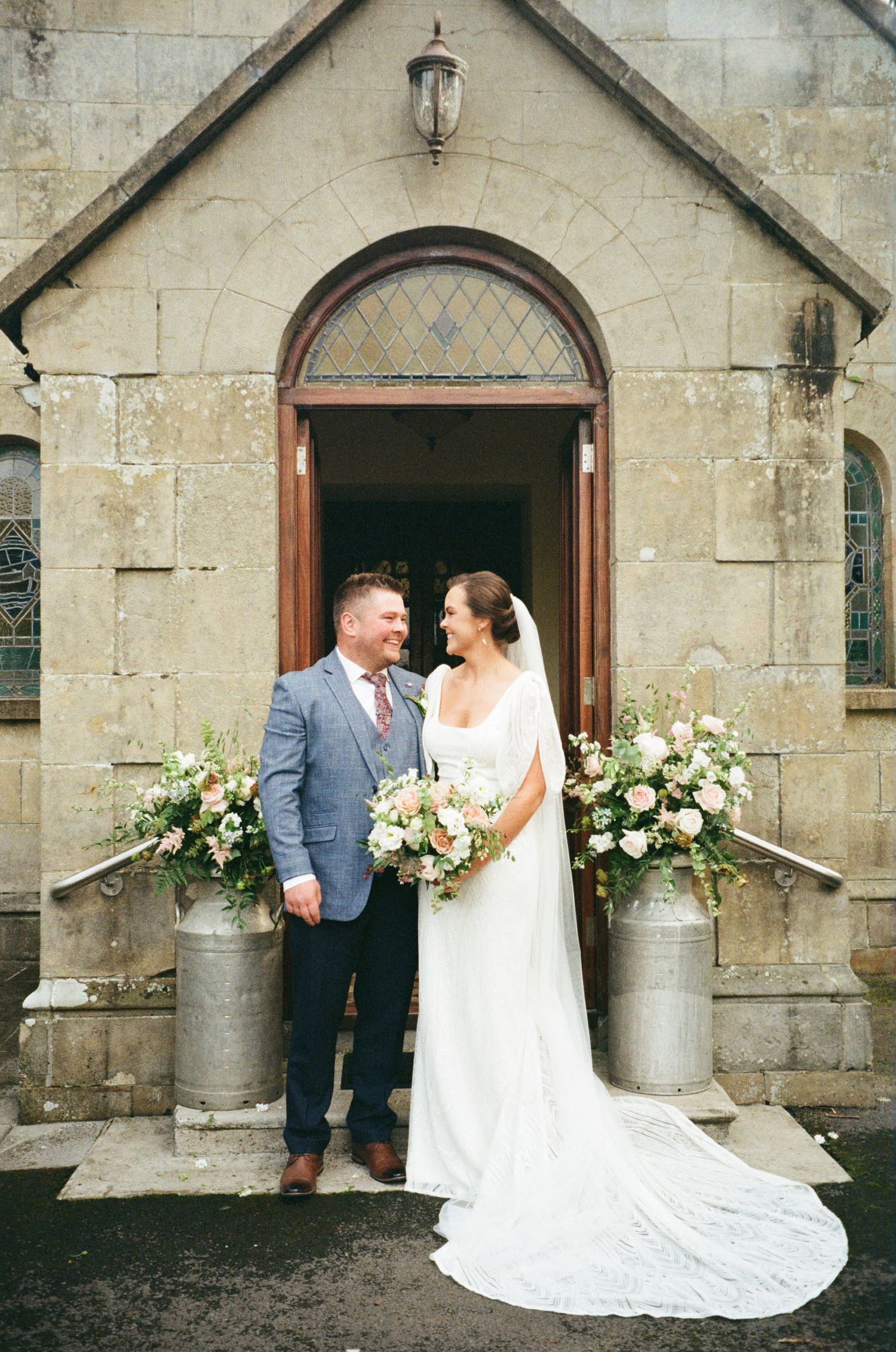 A romantic wedding couple shares tender moments outside a stone church with elegant floral arrangements in milk jugs.