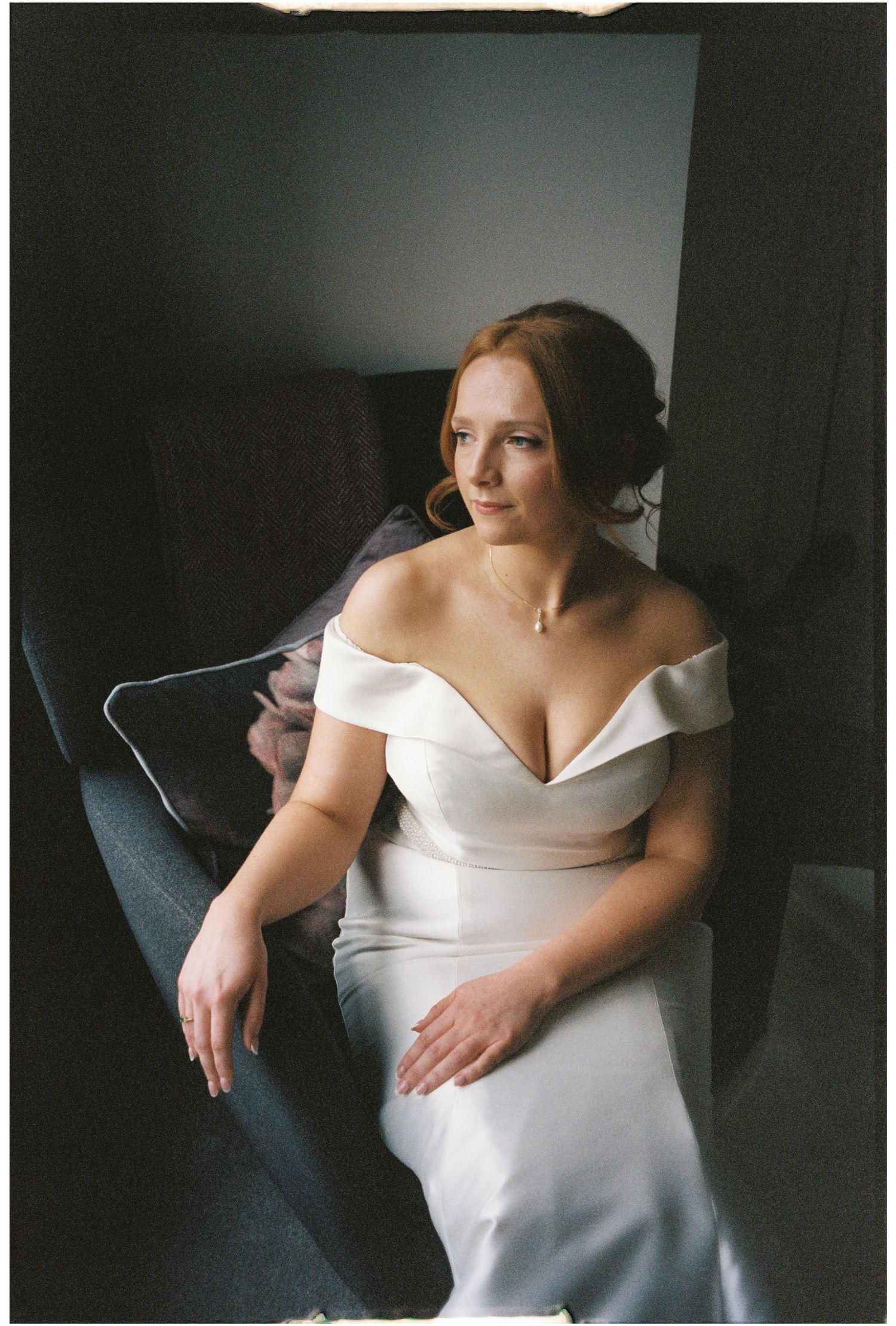 A figure in a sleek white off-shoulder wedding dress poses elegantly on a dark gray couch in moody lighting.