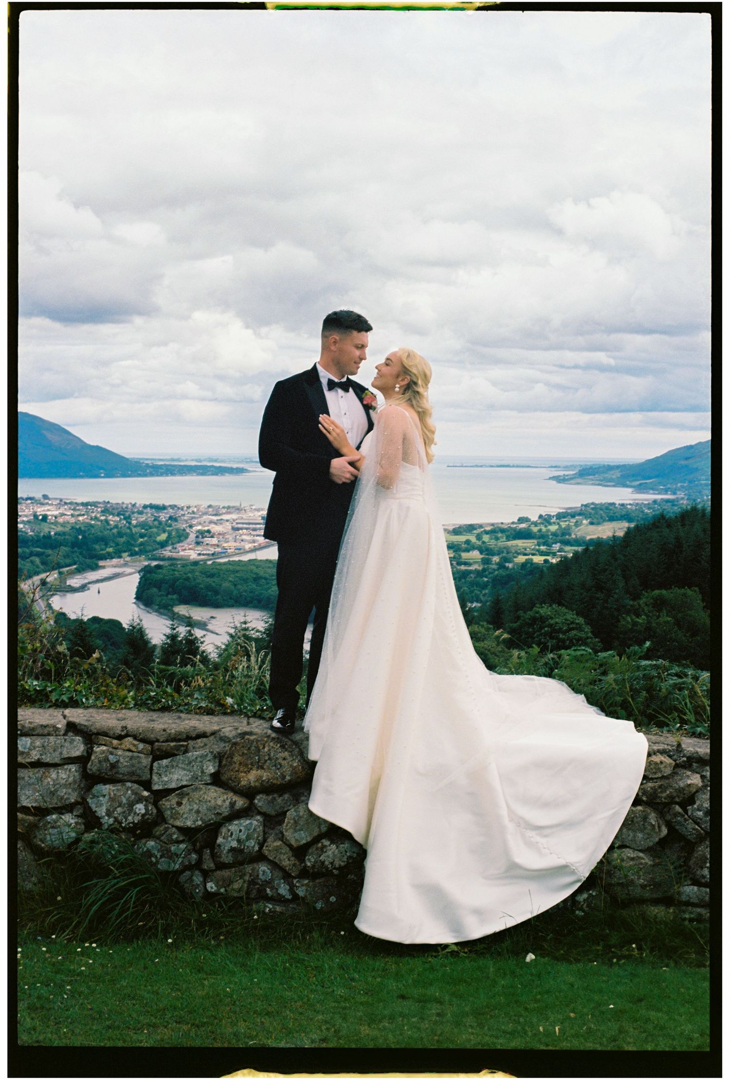 A wedding couple poses against a stone wall overlooking a scenic mountain valley and city landscape on an overcast day.
