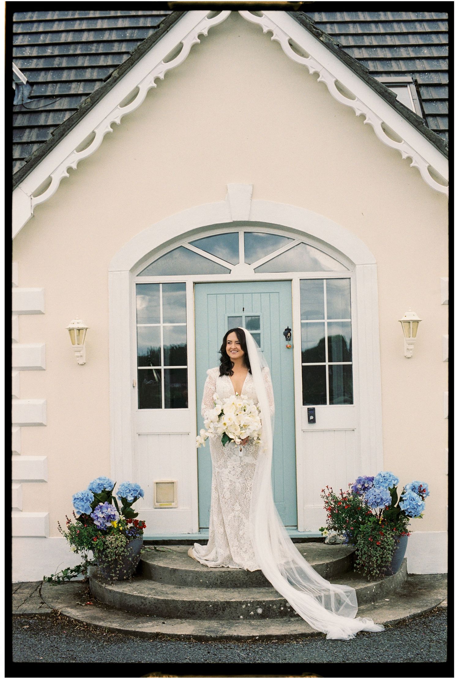 A white cottage-style house with blue door and hydrangea bushes serves as backdrop for bridal portraits.