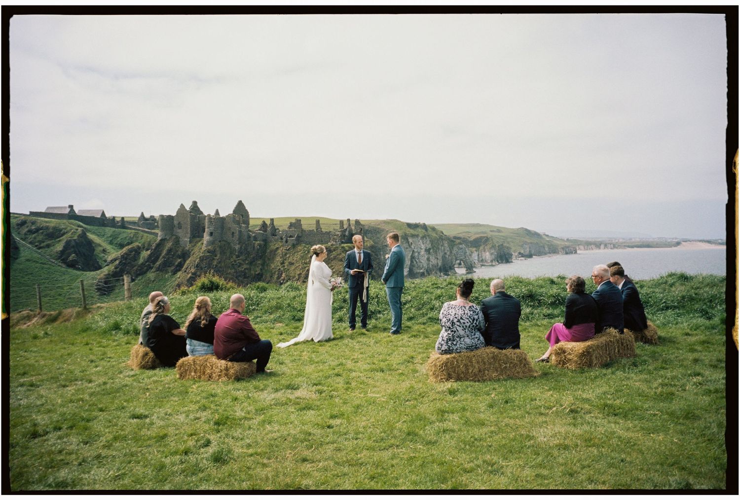 A group of people sit on a grassy hillside overlooking a scenic coastal landscape on a cloudy day.