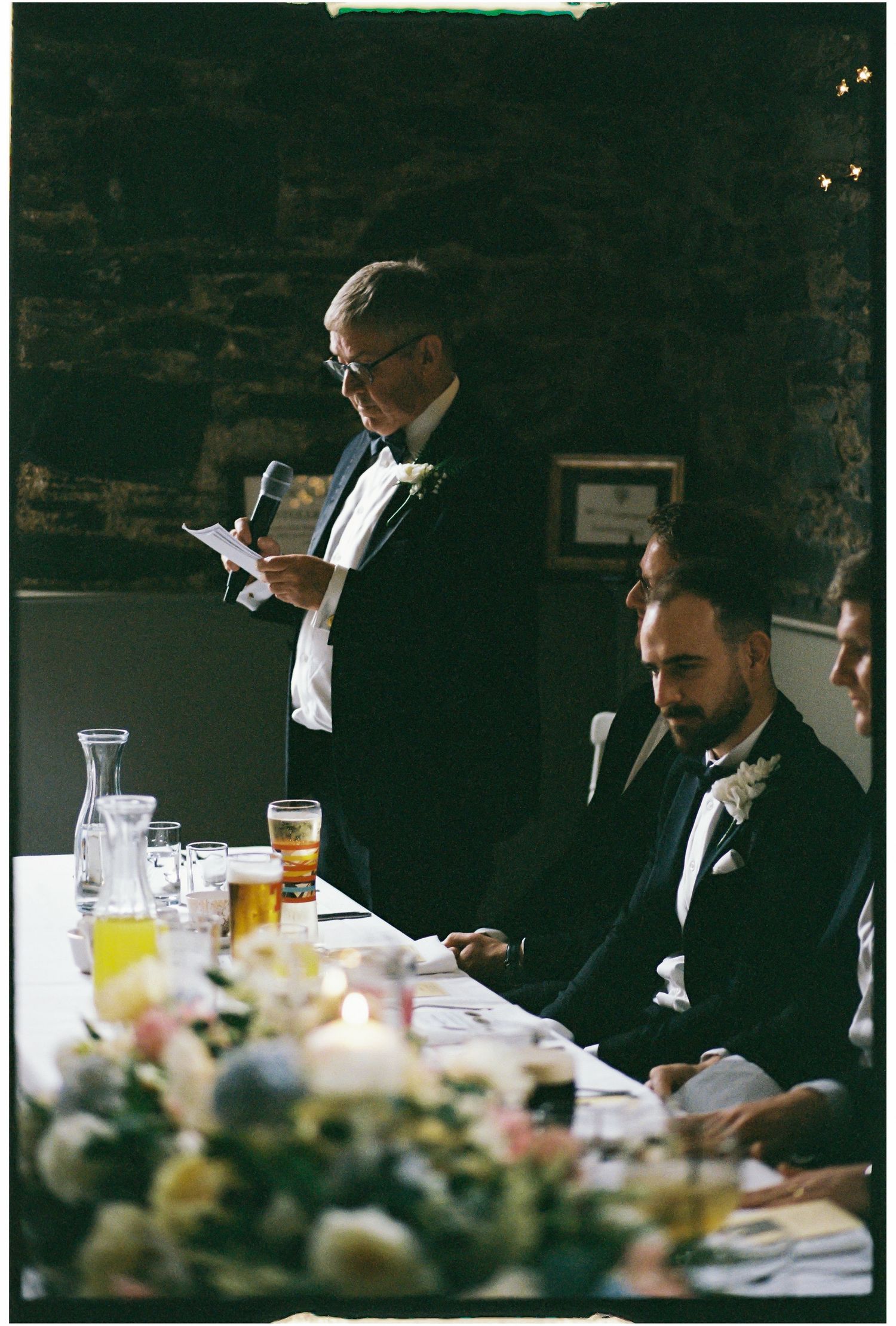 Wedding reception table with formally dressed guests enjoying drinks and conversation in dim lighting.