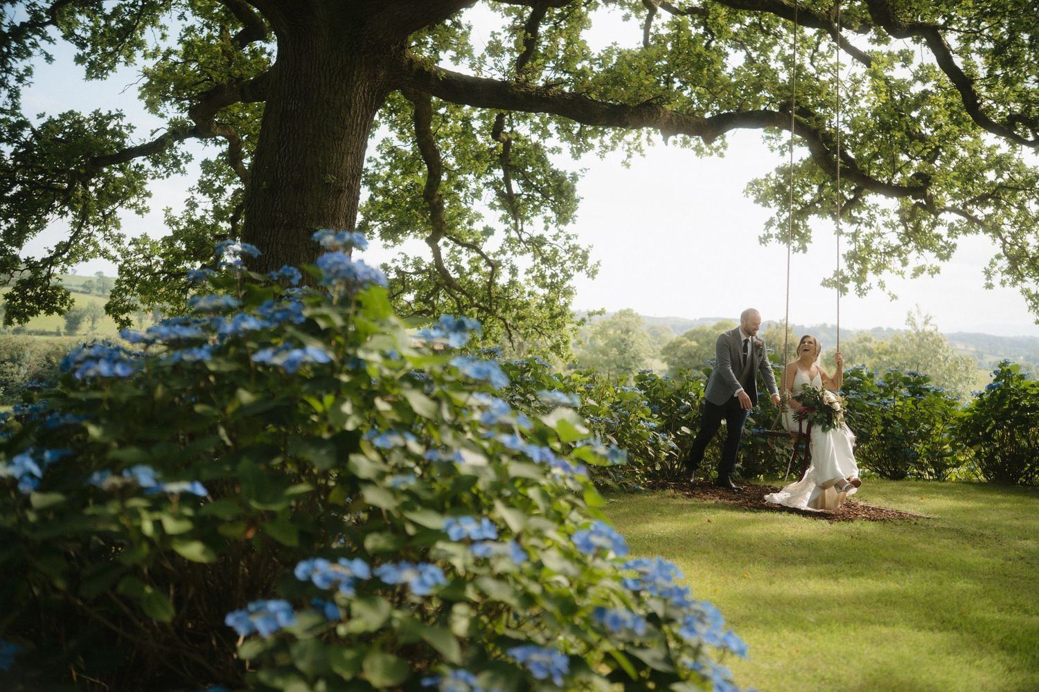 Wedding couple poses under lush green tree branches in a scenic outdoor setting.