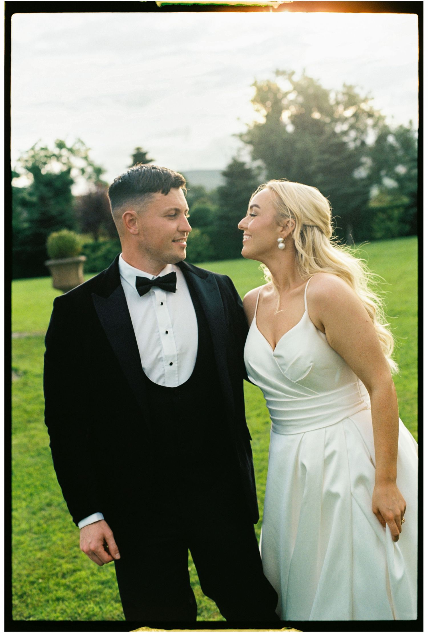Couple in formal wedding attire sharing a joyful moment together in a green outdoor setting at sunset.