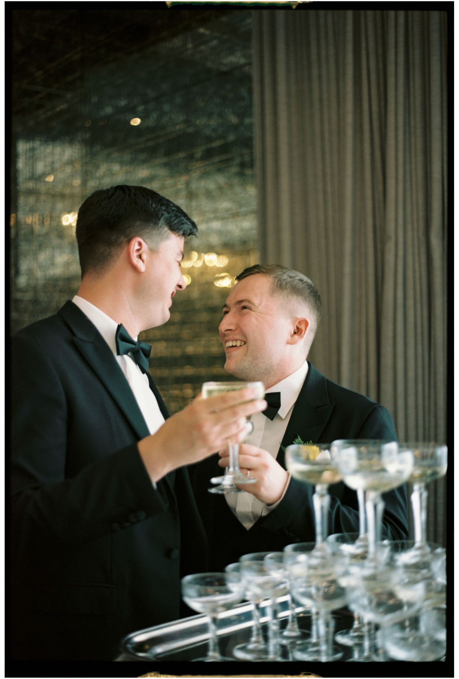 Two people in formal black attire adjusting bowties while smiling and sharing a moment together.