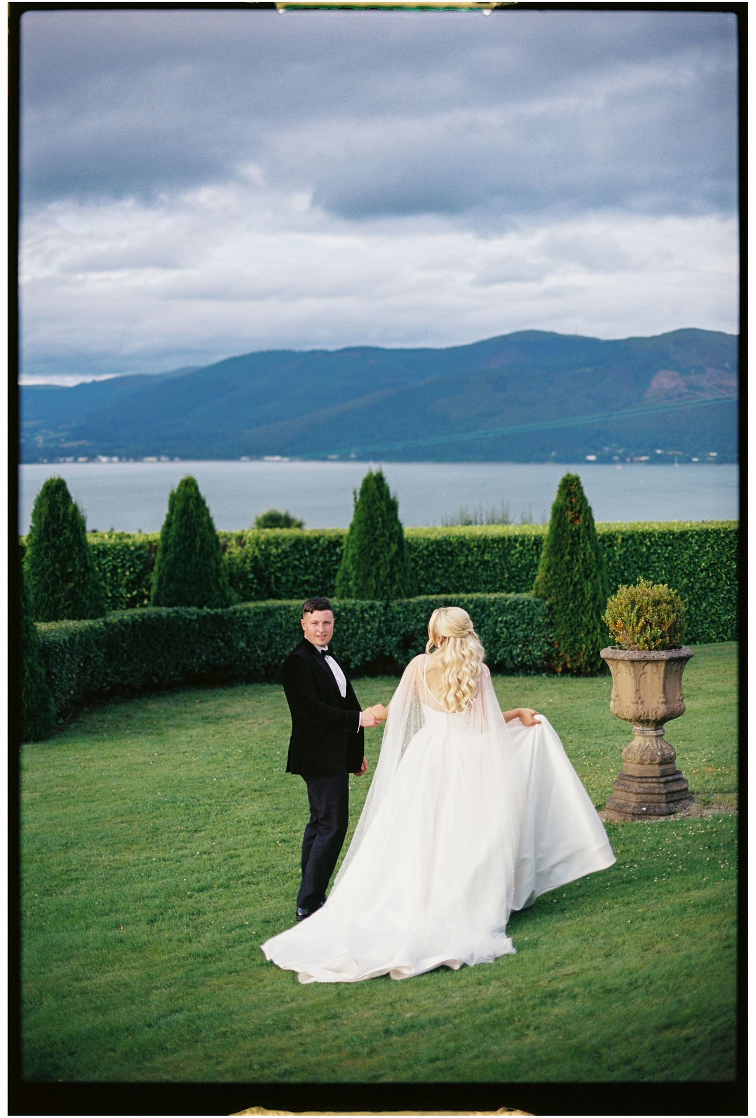 A wedding couple poses on a manicured lawn overlooking a scenic mountain lake view.