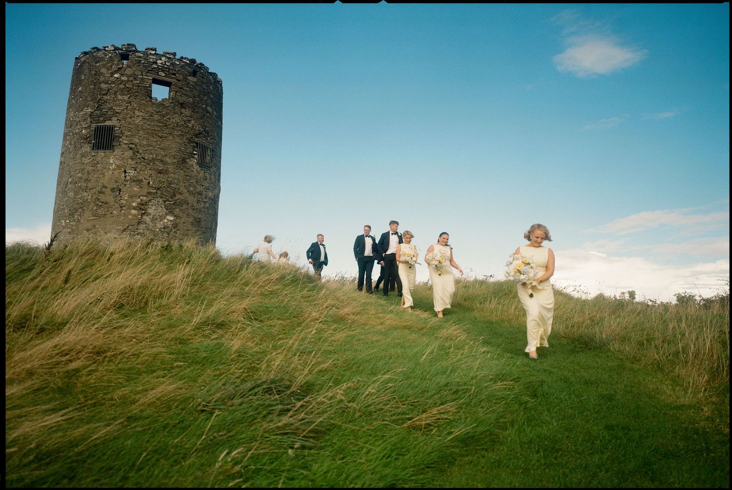 Wedding party walking along grassy hilltop path with ancient stone ruins in the background.