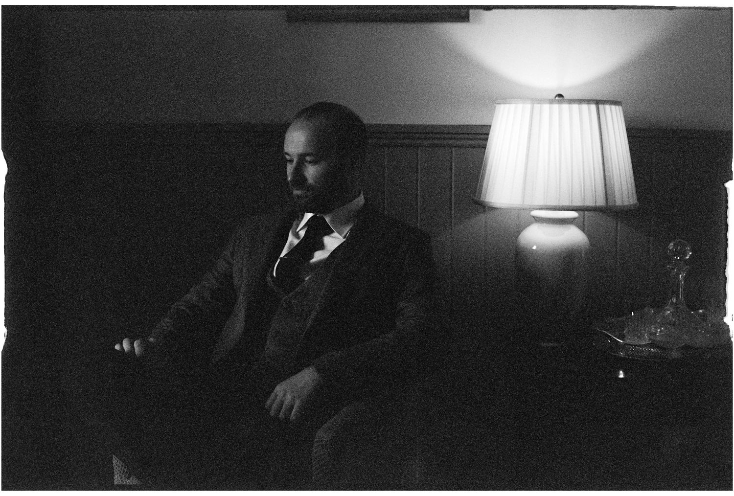A person sits in a dimly lit room with a table lamp casting moody film noir-style lighting in black and white.