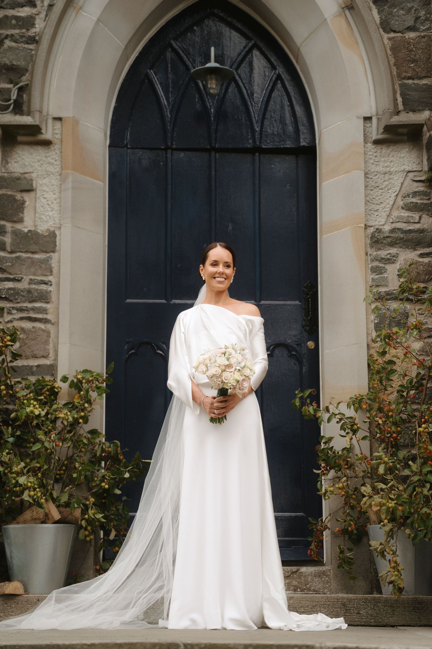 A bride in a long white wedding dress with a flowing cape stands in front of an arched church doorway.