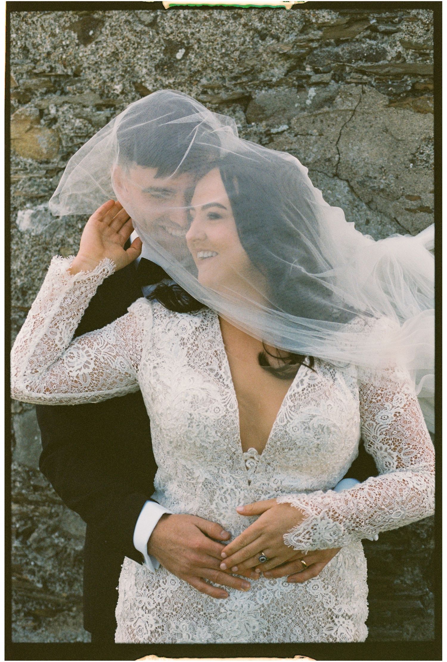 Romantic wedding portrait with lace dress and flowing veil against stone wall backdrop.