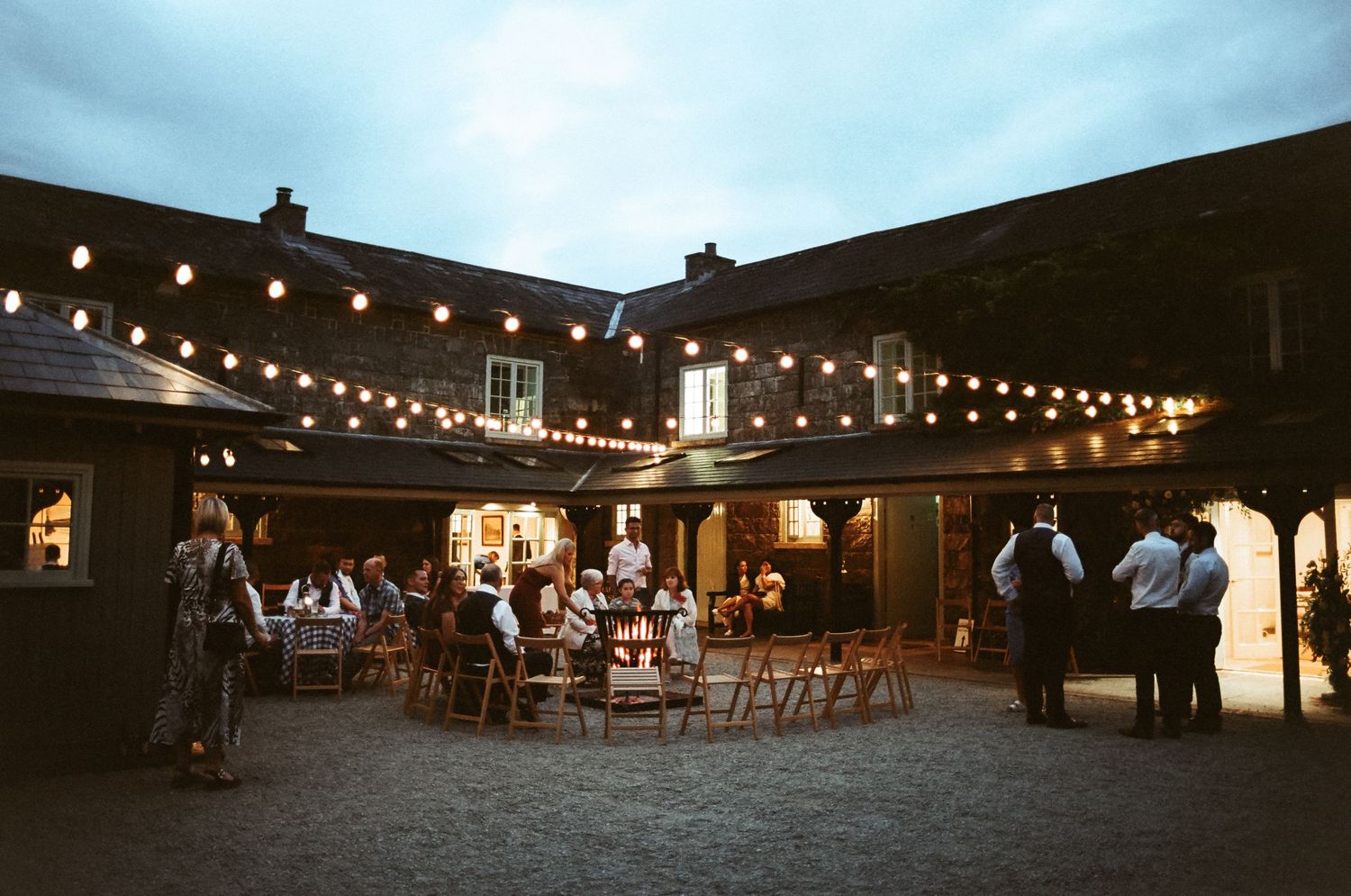 String lights illuminate a rustic barn venue at dusk during an outdoor wedding reception.