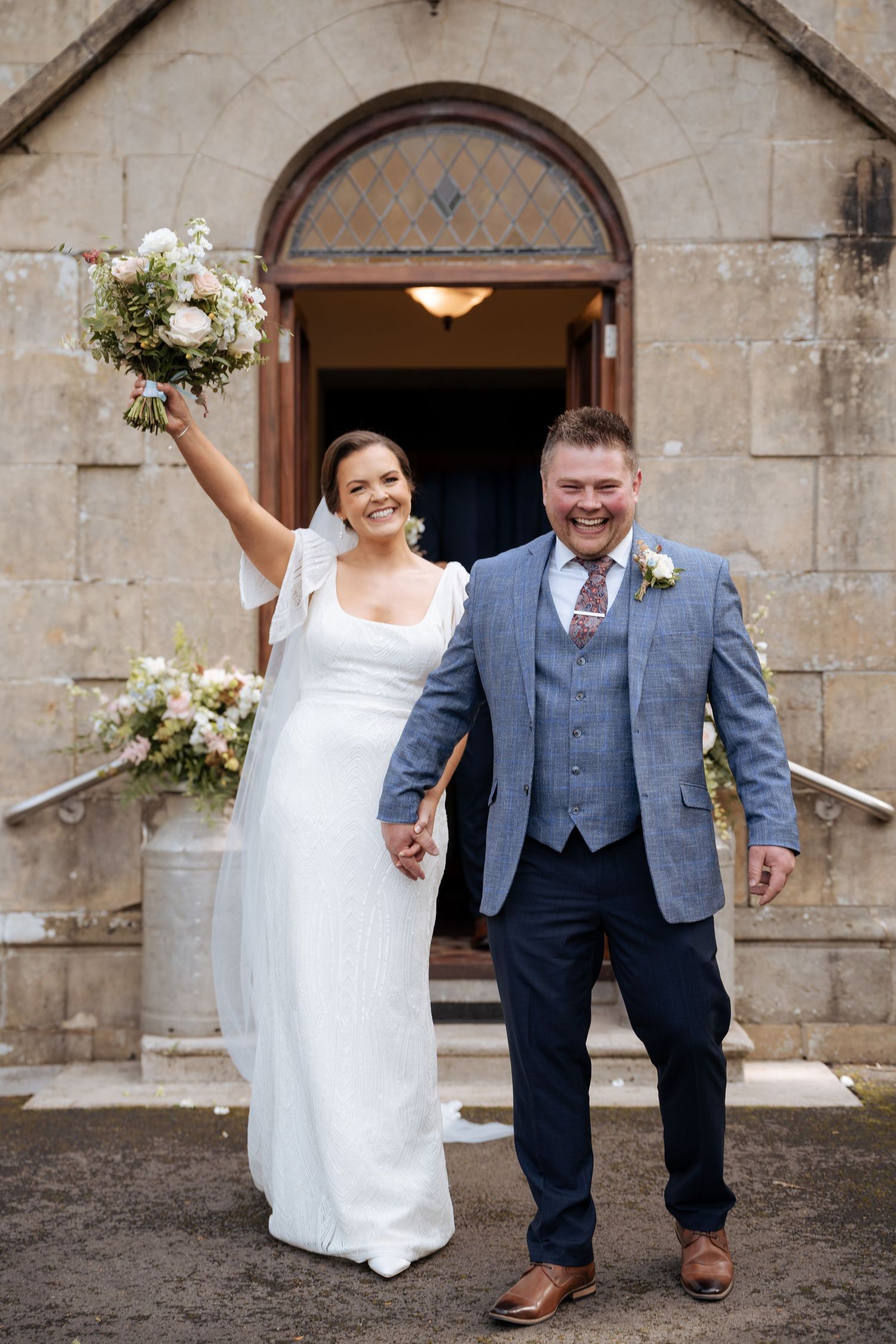 Newlyweds celebrate while exiting stone church building with bouquet raised in triumph.