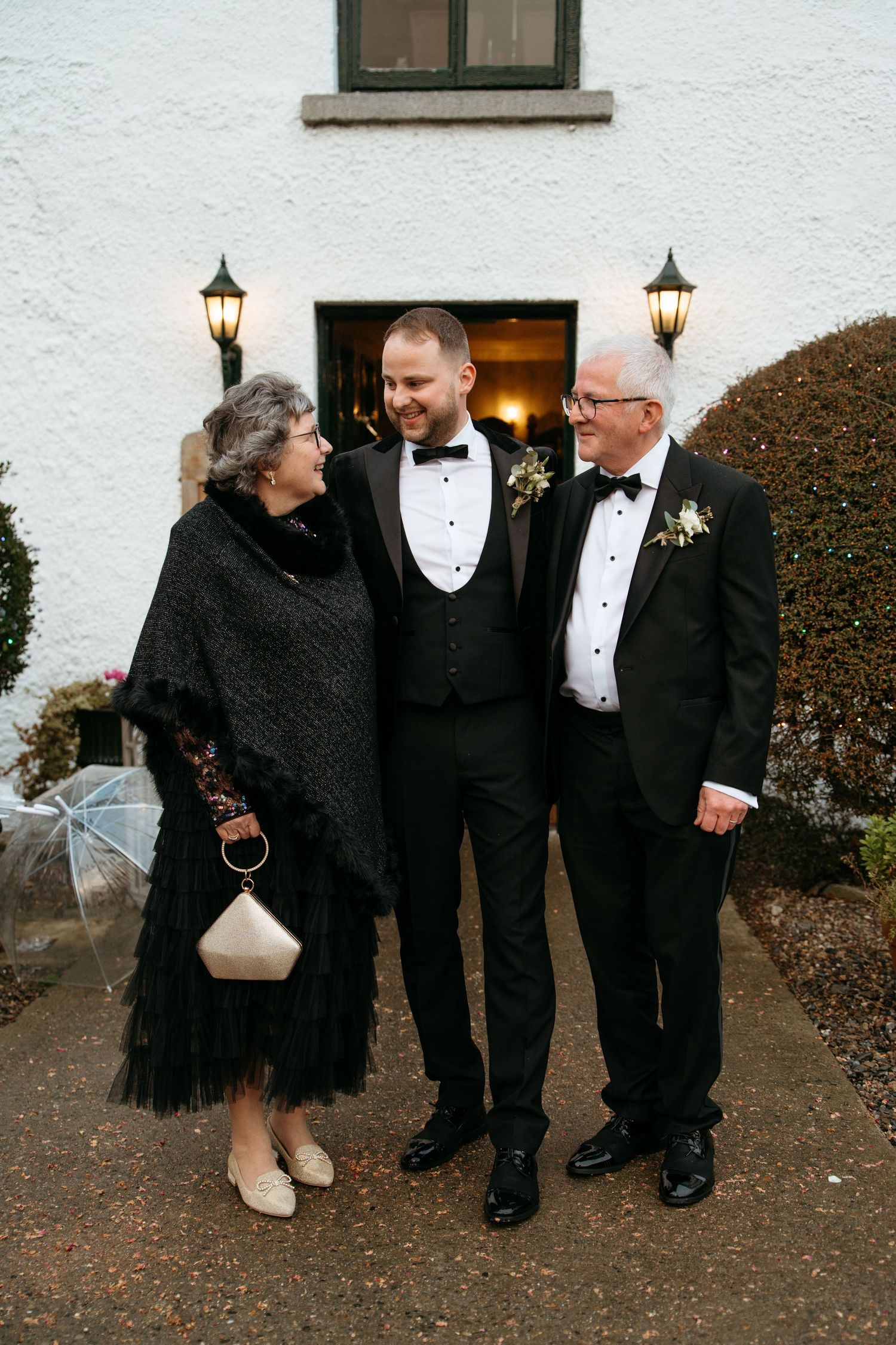 Wedding guests in formal black attire laughing together outside a white building.