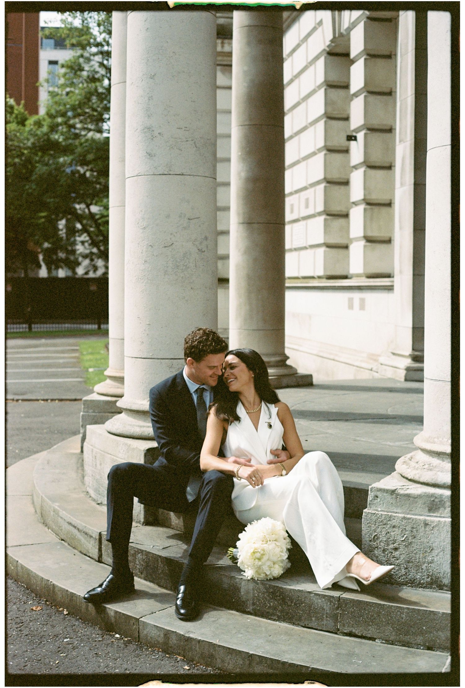 A couple in wedding attire sits intimately on stone steps between classical white columns in a romantic outdoor setting.
