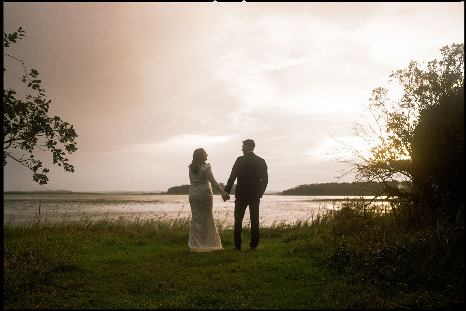 A couple stands silhouetted against a sunset over a lake, surrounded by natural scenery.