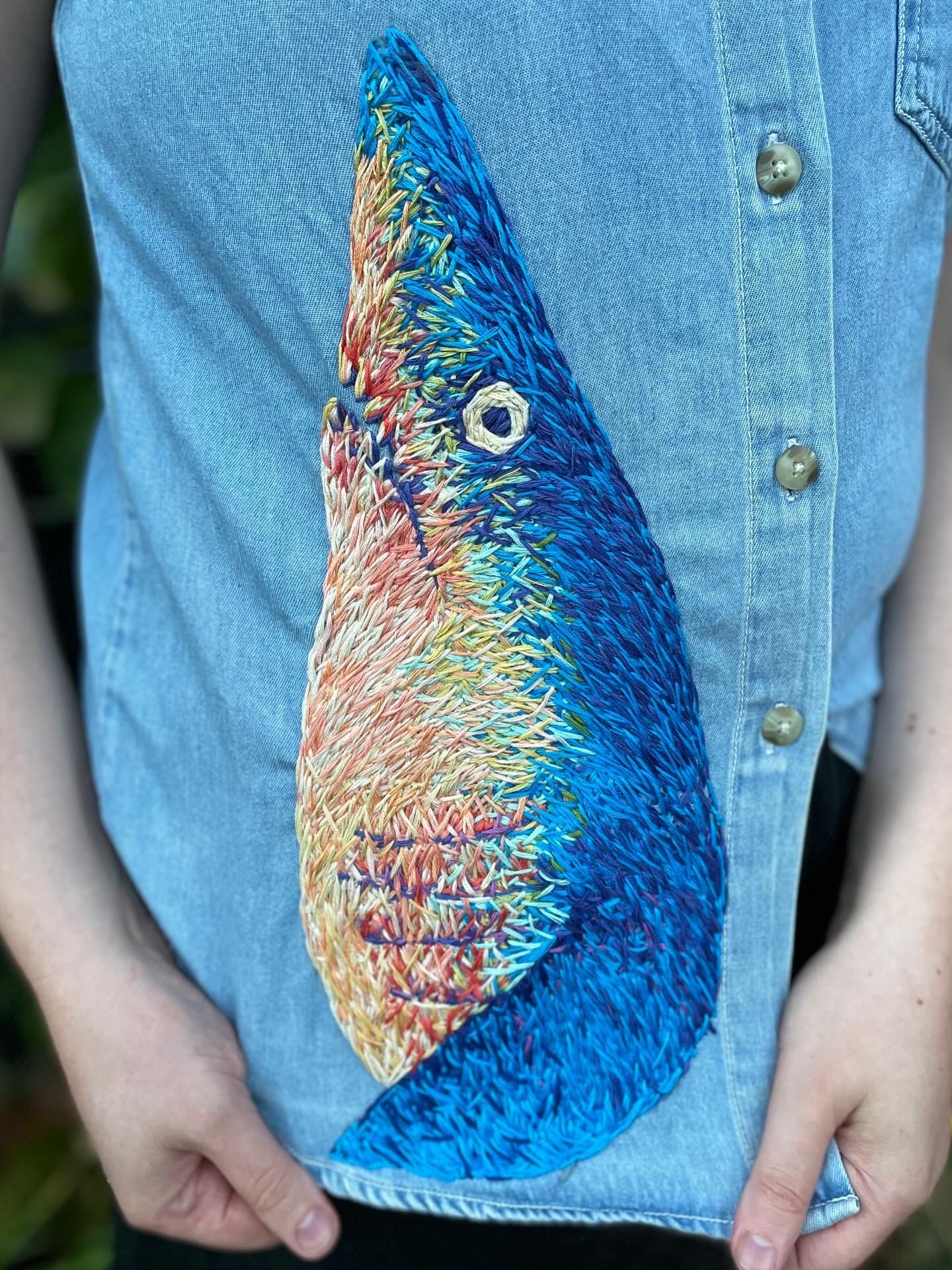 Detailed colorful embroidery of a mako shark stitched onto a denim shirt with vibrant blue, orange and yellow threads.