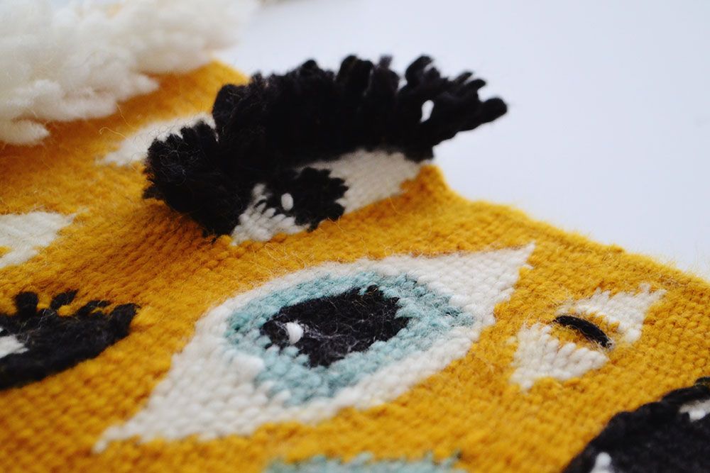 Close-up detail of an orange woven textile with eye motifs and black yarn eyelashes.
