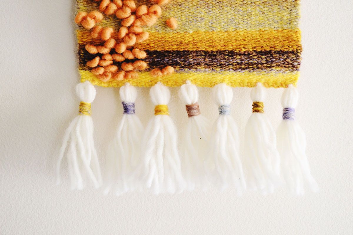Close-up of a woven wall tapestry with white tassels and orange, yellow, and brown yarn.