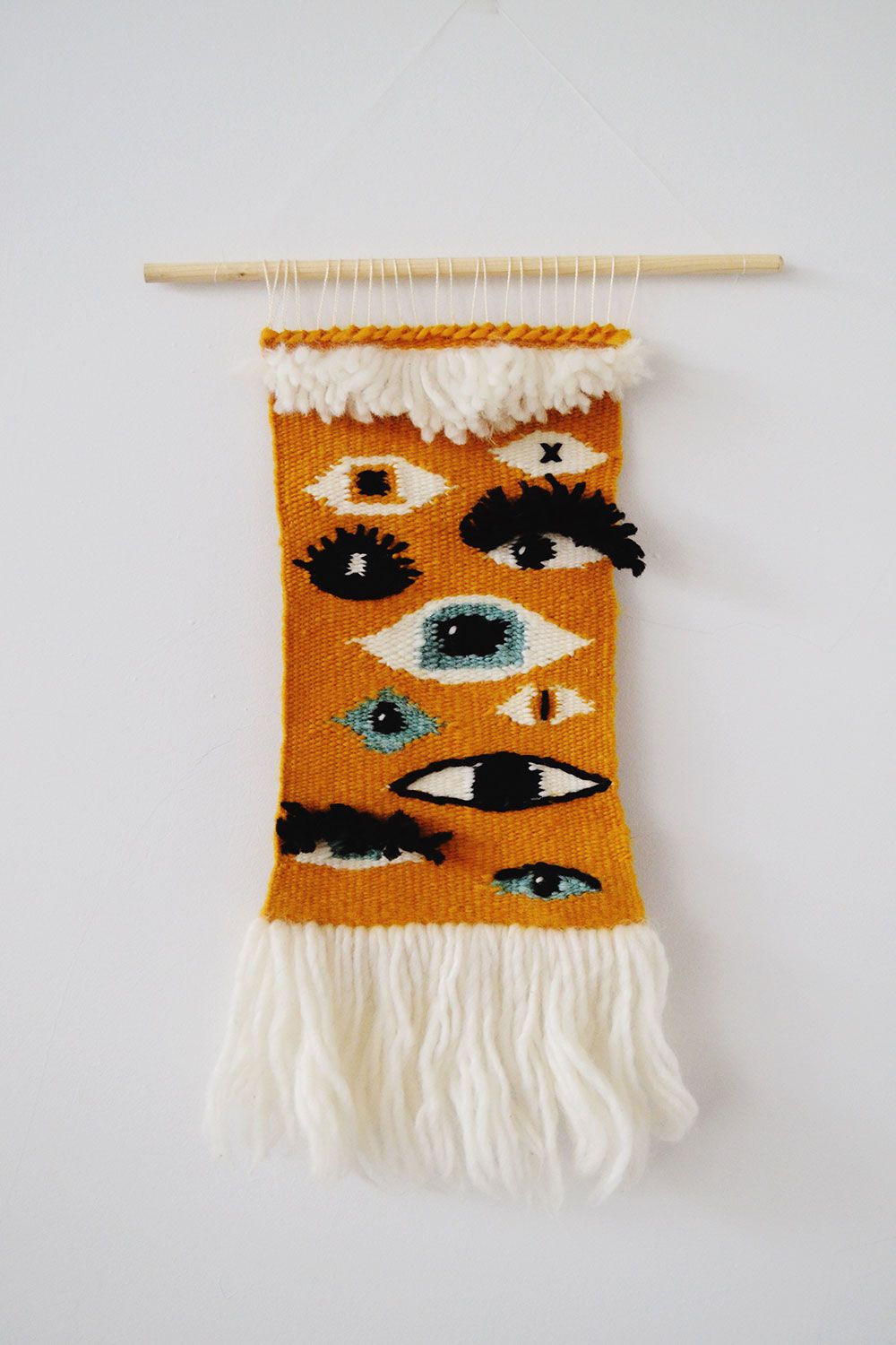 Orange woven wall tapestry featuring multiple eye designs with white fringe on a wooden dowel.