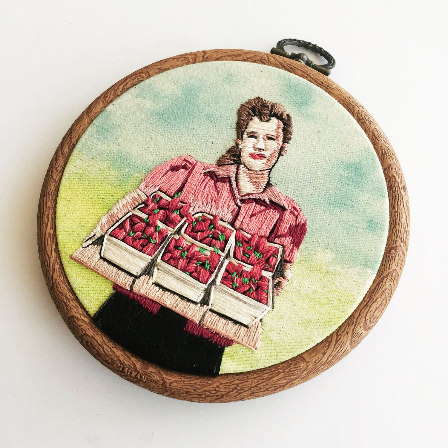 Embroidery hoop artwork depicting a woman holding trays of strawberries with watercolor background in wooden frame.
