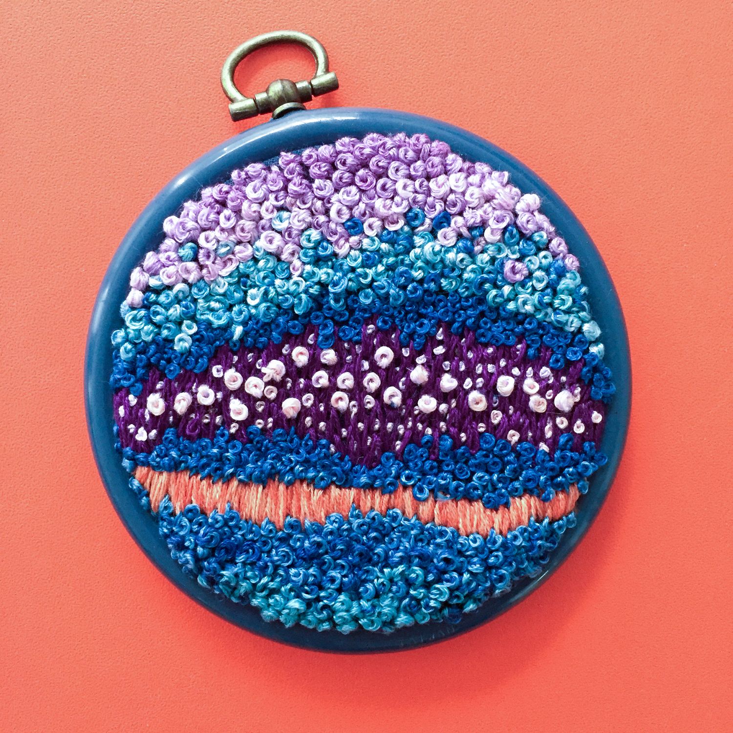 Circular embroidery hoop featuring intricate French knots in purple, blue and orange wave pattern on red background.
