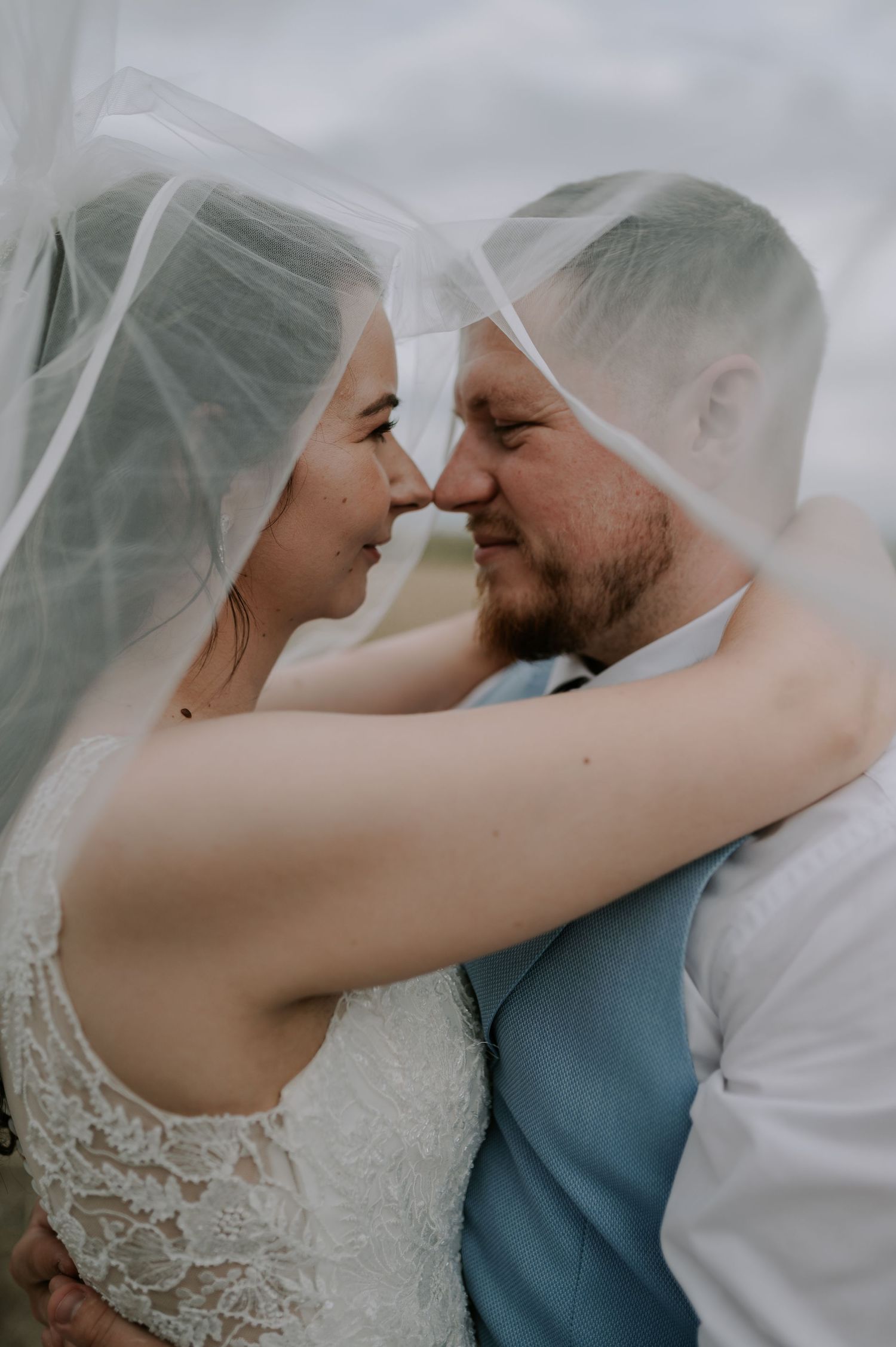 A romantic moment captured between newlyweds under a flowing white veil against an overcast sky.