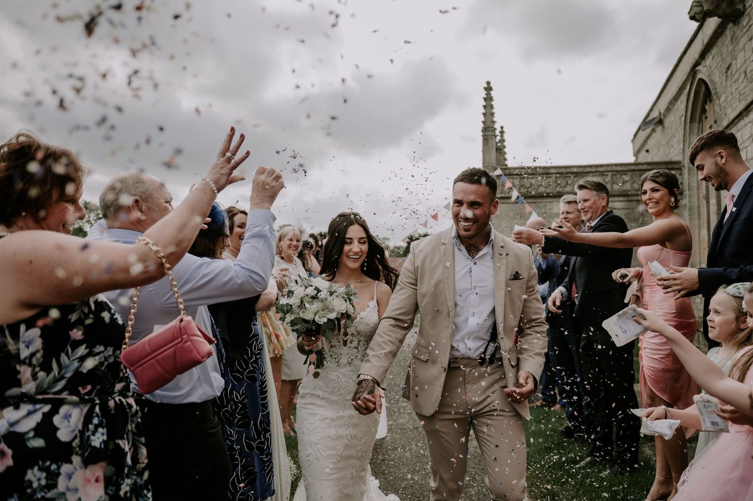 Wedding celebration guests toss rose petals and confetti over a joyful couple exiting their ceremony together.