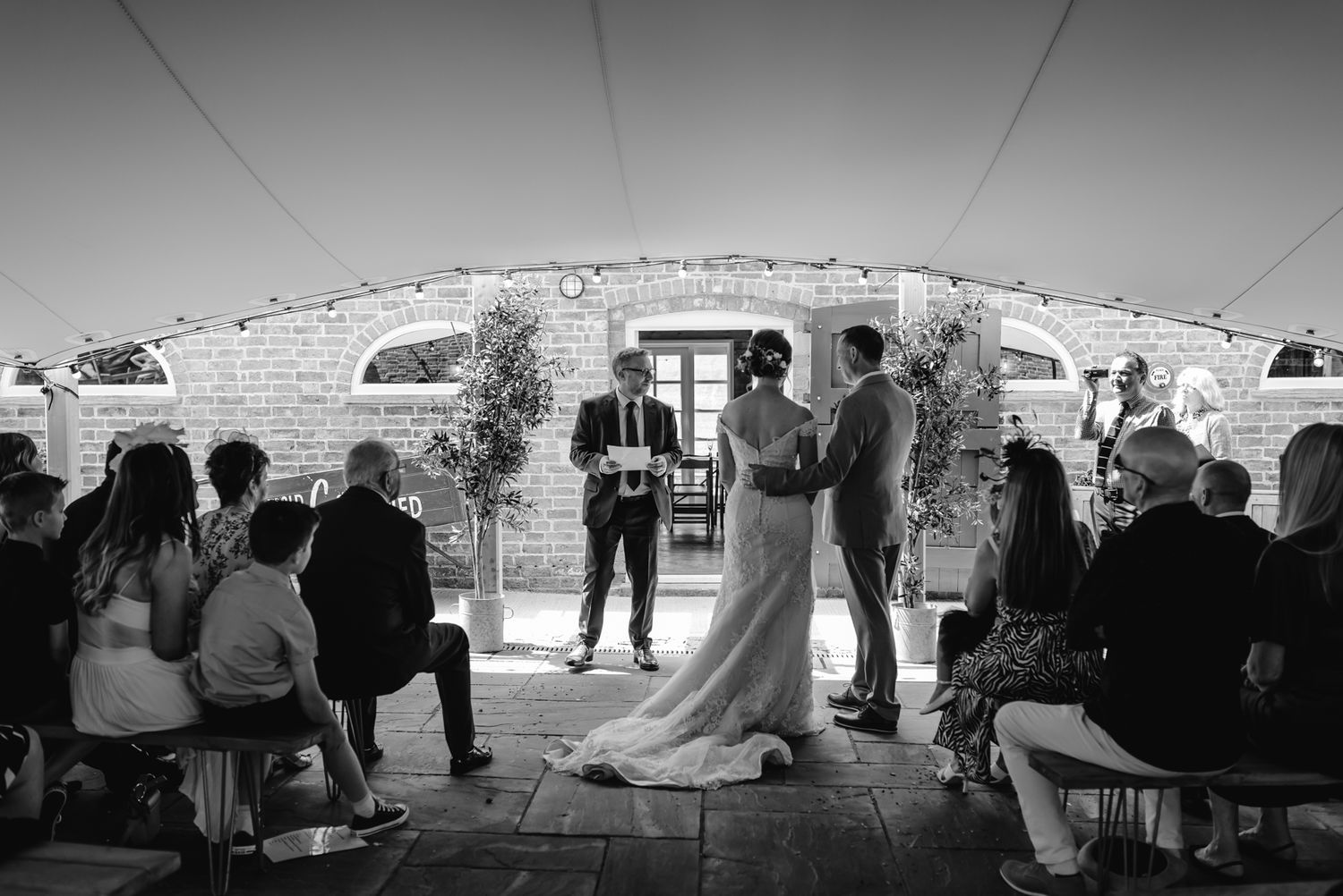 Wedding ceremony taking place under a stone archway with guests seated on both sides in black and white.