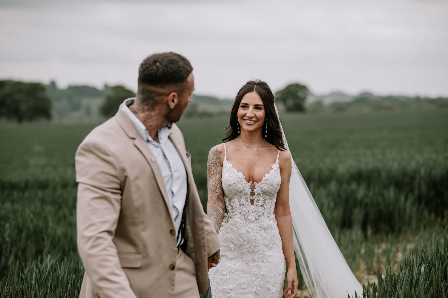 A romantic outdoor wedding scene shows a couple holding hands in a lush green field during golden hour.