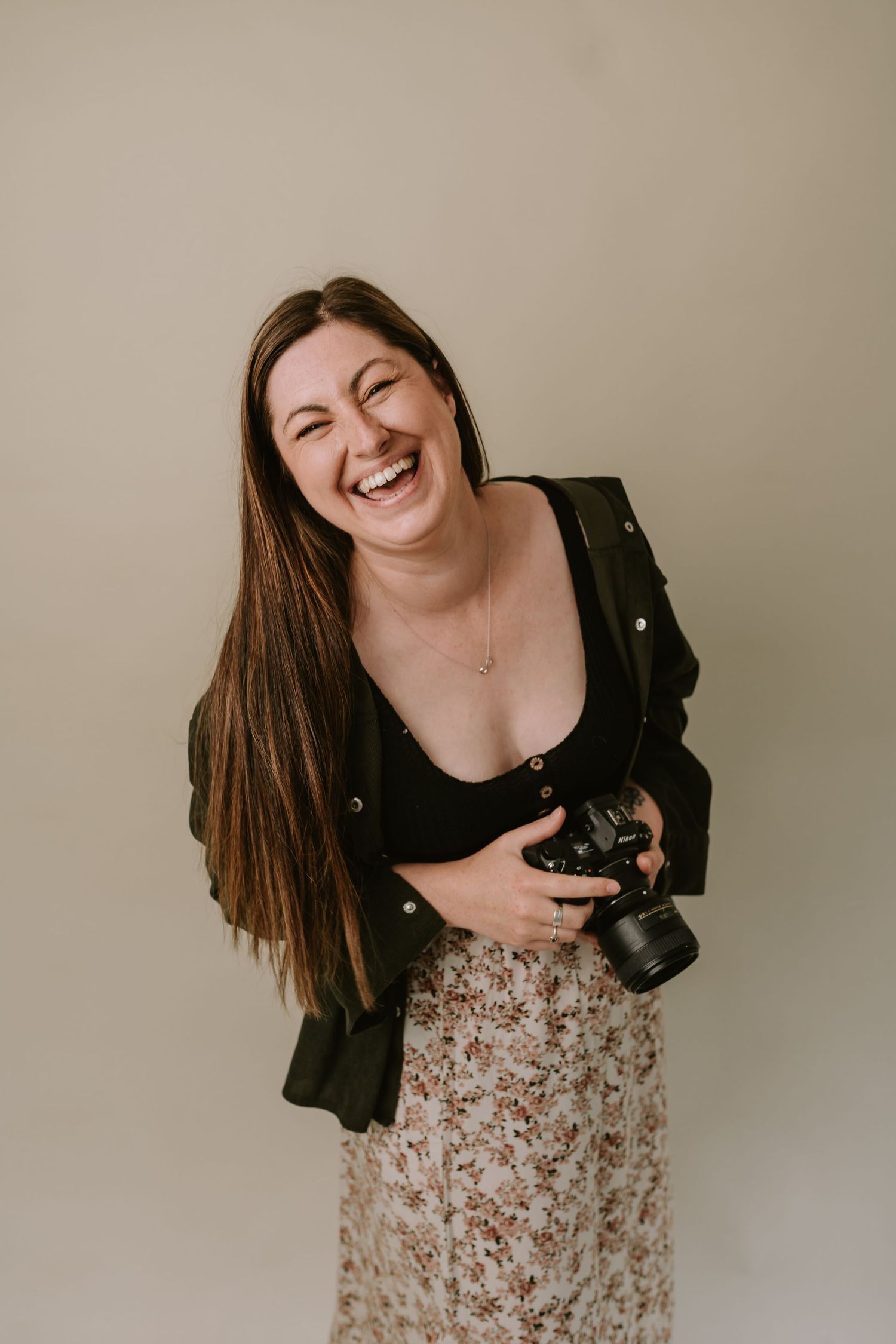 A photographer in a black jacket and floral skirt laughs joyfully while holding a professional camera against a neutral backdrop.