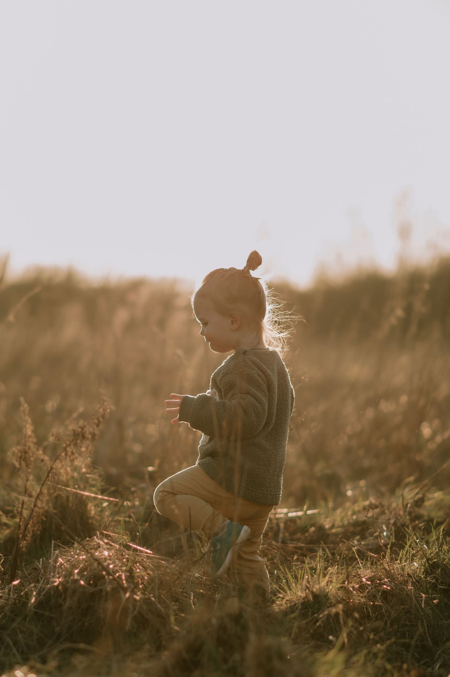Toddler in olive green outfit playing in golden sunlit field during sunset, hair blowing in breeze.