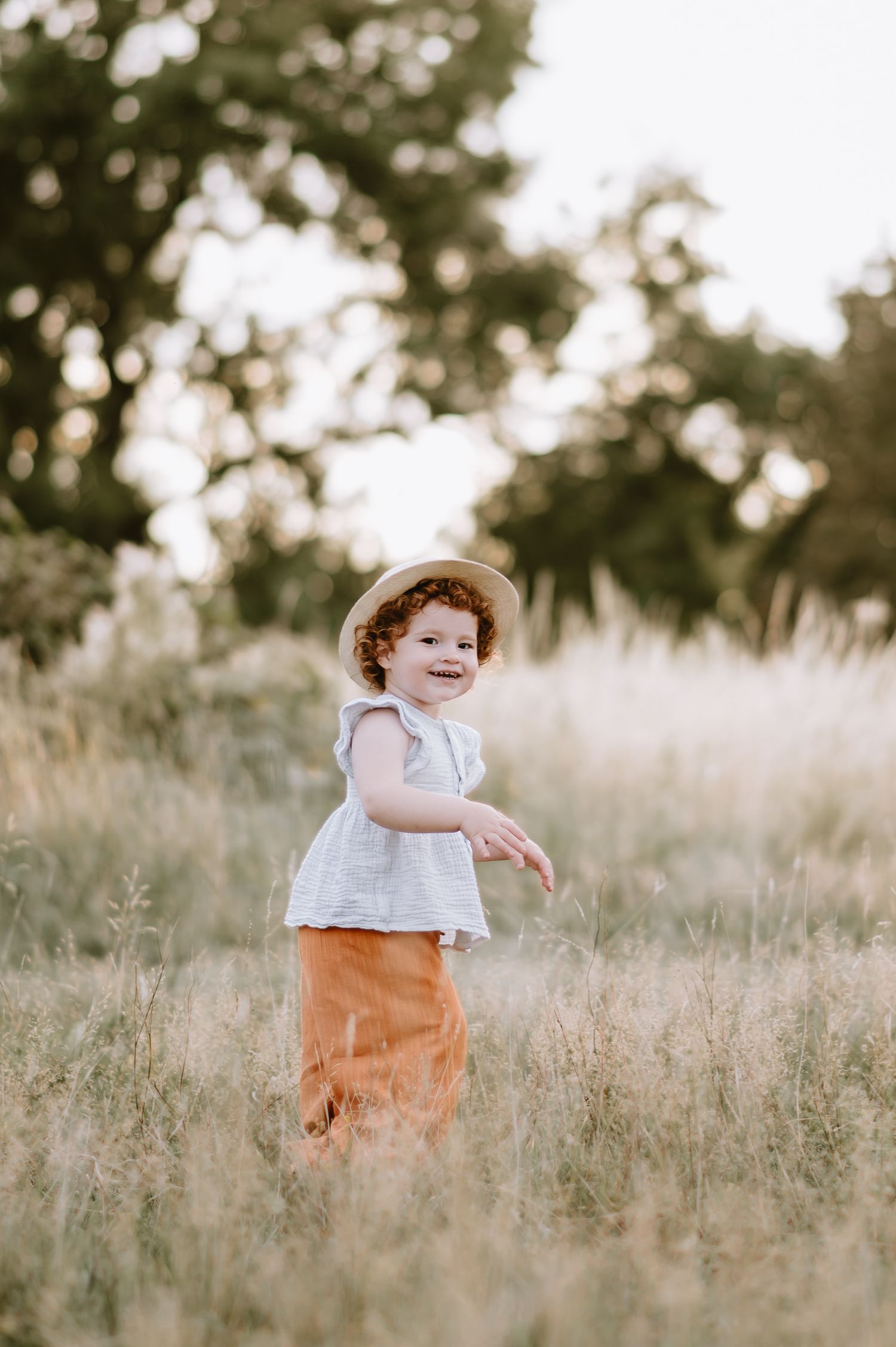 Curly-haired toddler in straw hat and white top smiling in golden meadow field during sunset.