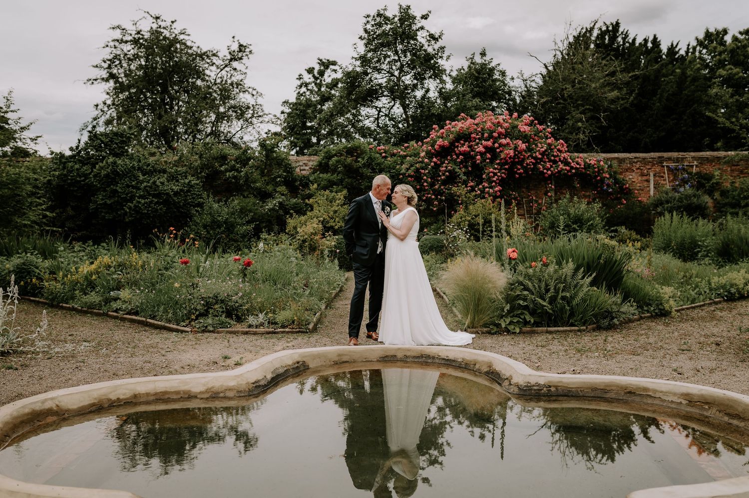 Bride and groom standing by a reflective garden pond surrounded by lush greenery and blooming flowers.