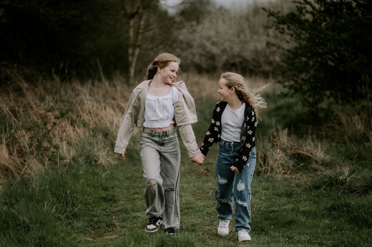 Two young girls holding hands and smiling while walking together through a lush green meadow path.