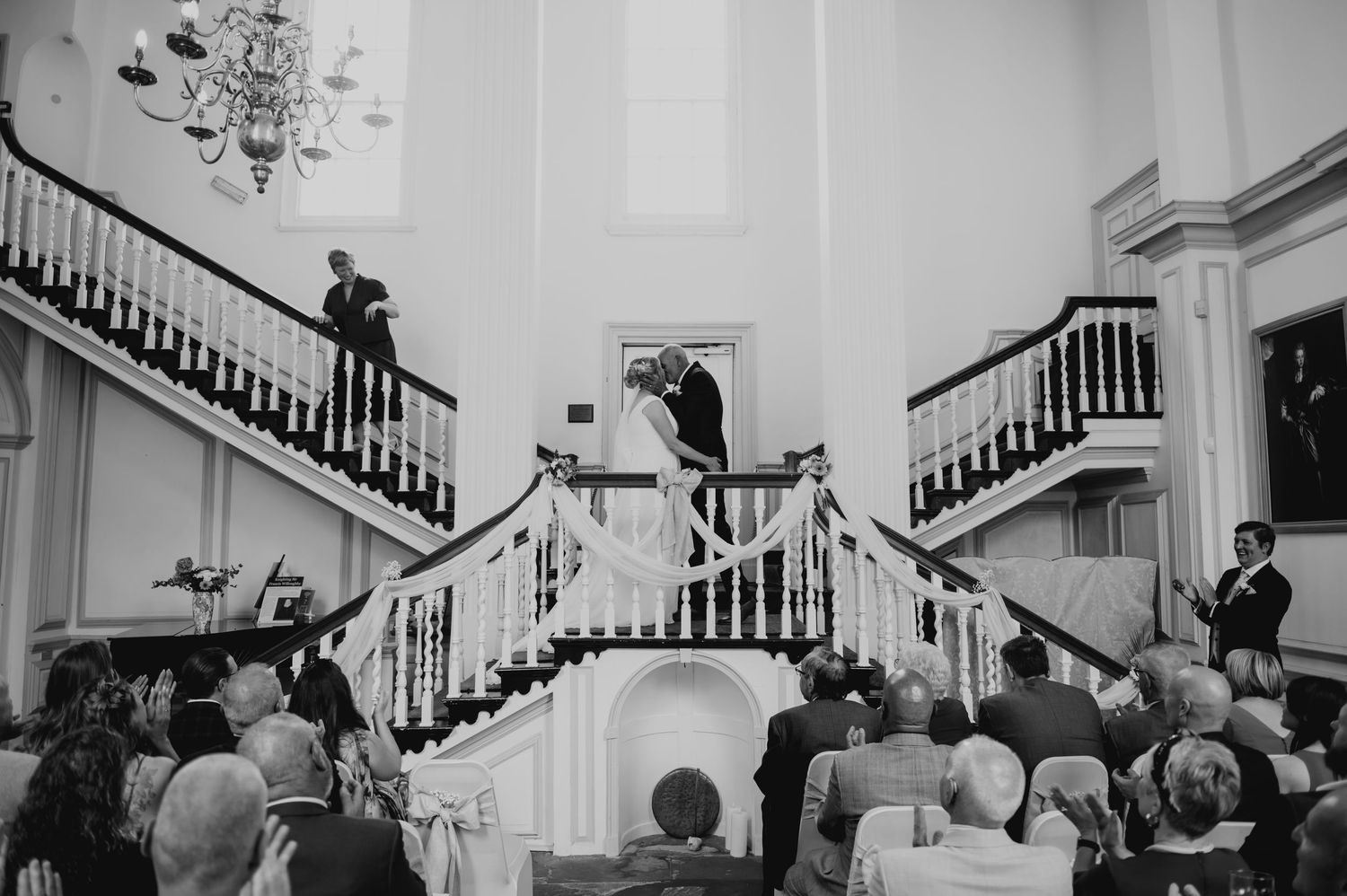 Black and white wedding ceremony photo of bride and groom kissing on grand staircase before seated guests.