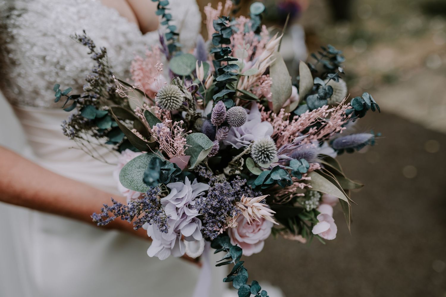 A delicate bridal bouquet featuring soft pink roses, lavender flowers, and eucalyptus greenery held against a blurred background.