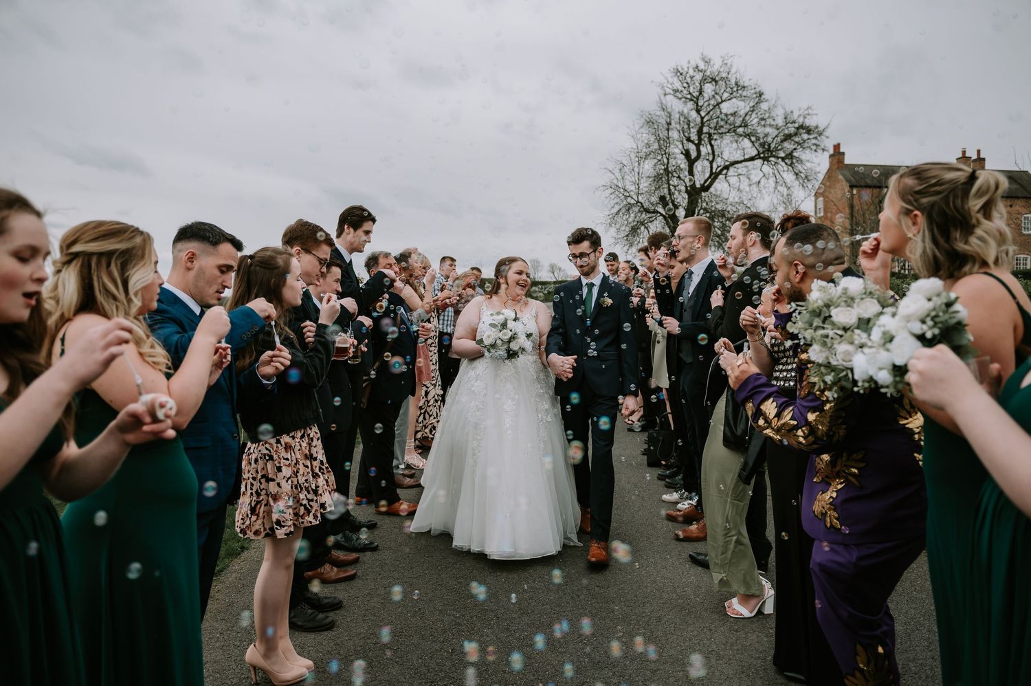 Wedding guests in formal attire form an aisle on a cloudy day as they blow bubbles during an outdoor celebration.