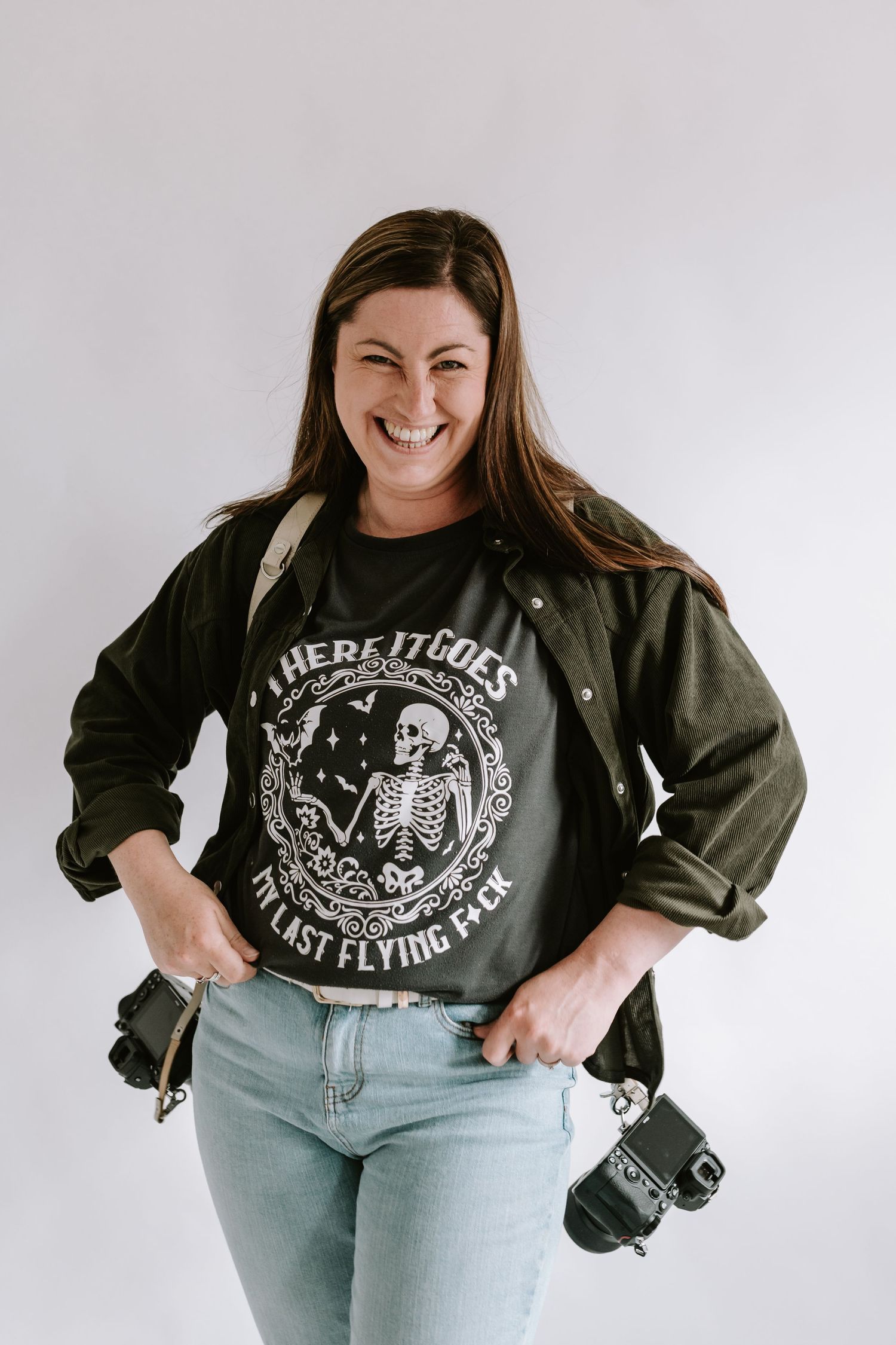 A person wearing a black t-shirt with skeleton graphic and light jeans holds a camera while smiling against a white background.