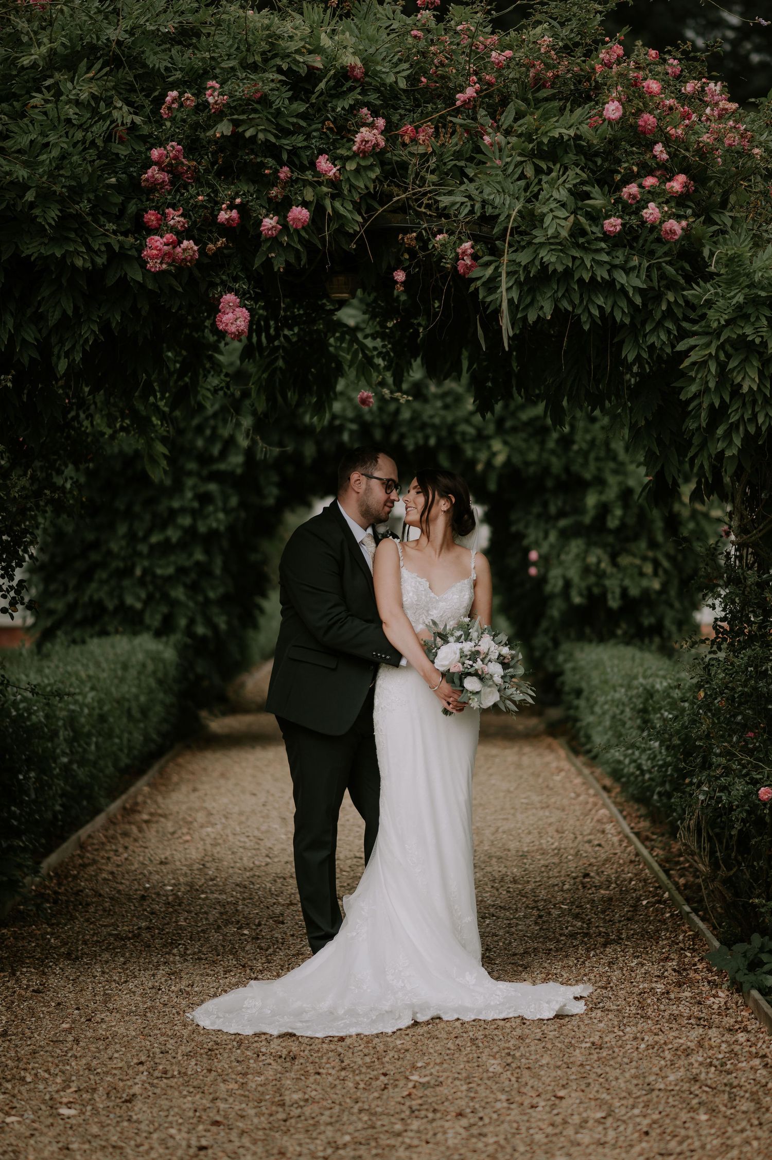 Bride and groom with the bride’s back to the groom’s chest, under an archway of red roses and greenery – Melton Mowbray wedding.