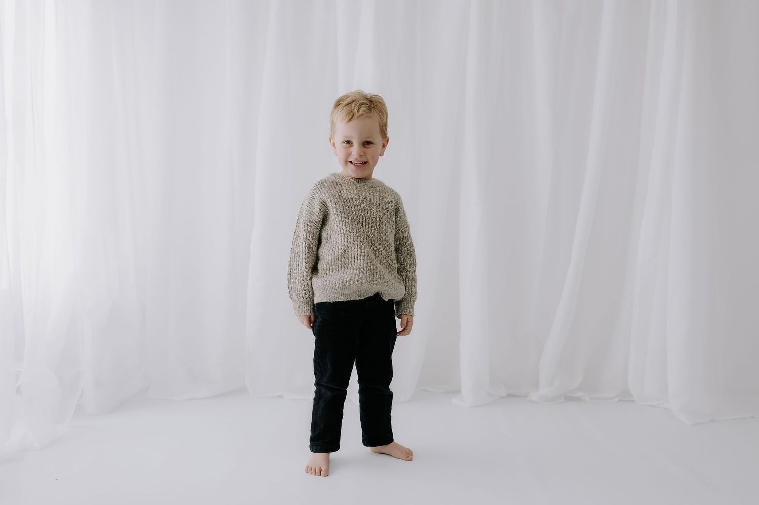 Boy in grey jumper and jeans stands smiling in a white studio. Melton Mowbray children’s photoshoot