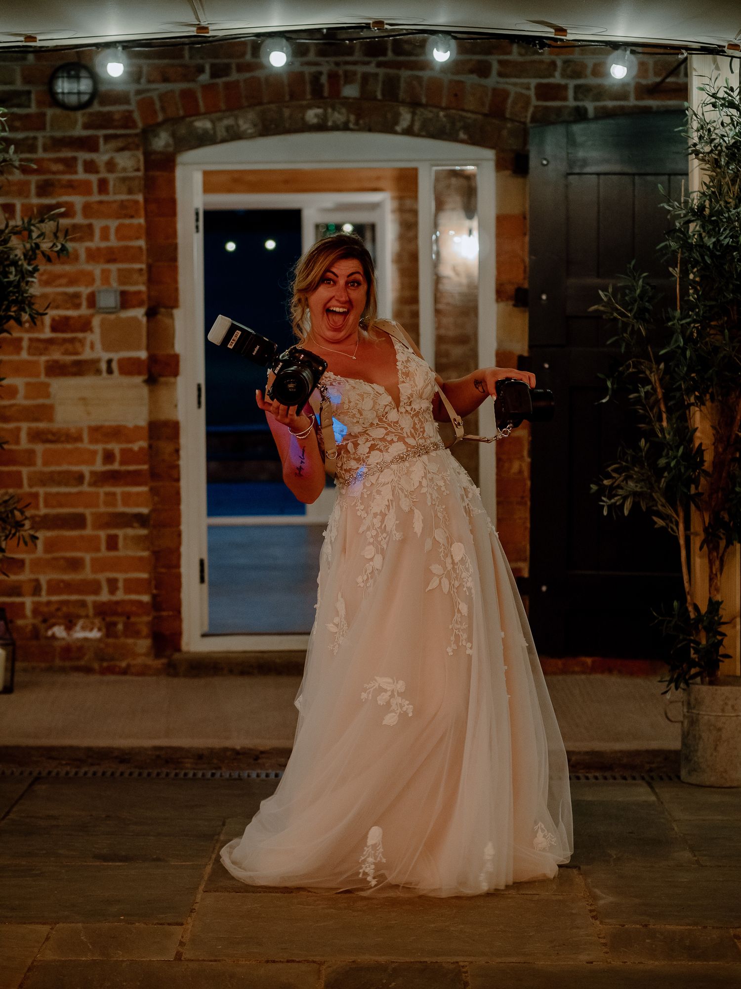 Bride holding a camera and posing playfully during the wedding reception, with warm ambient lighting in the background.