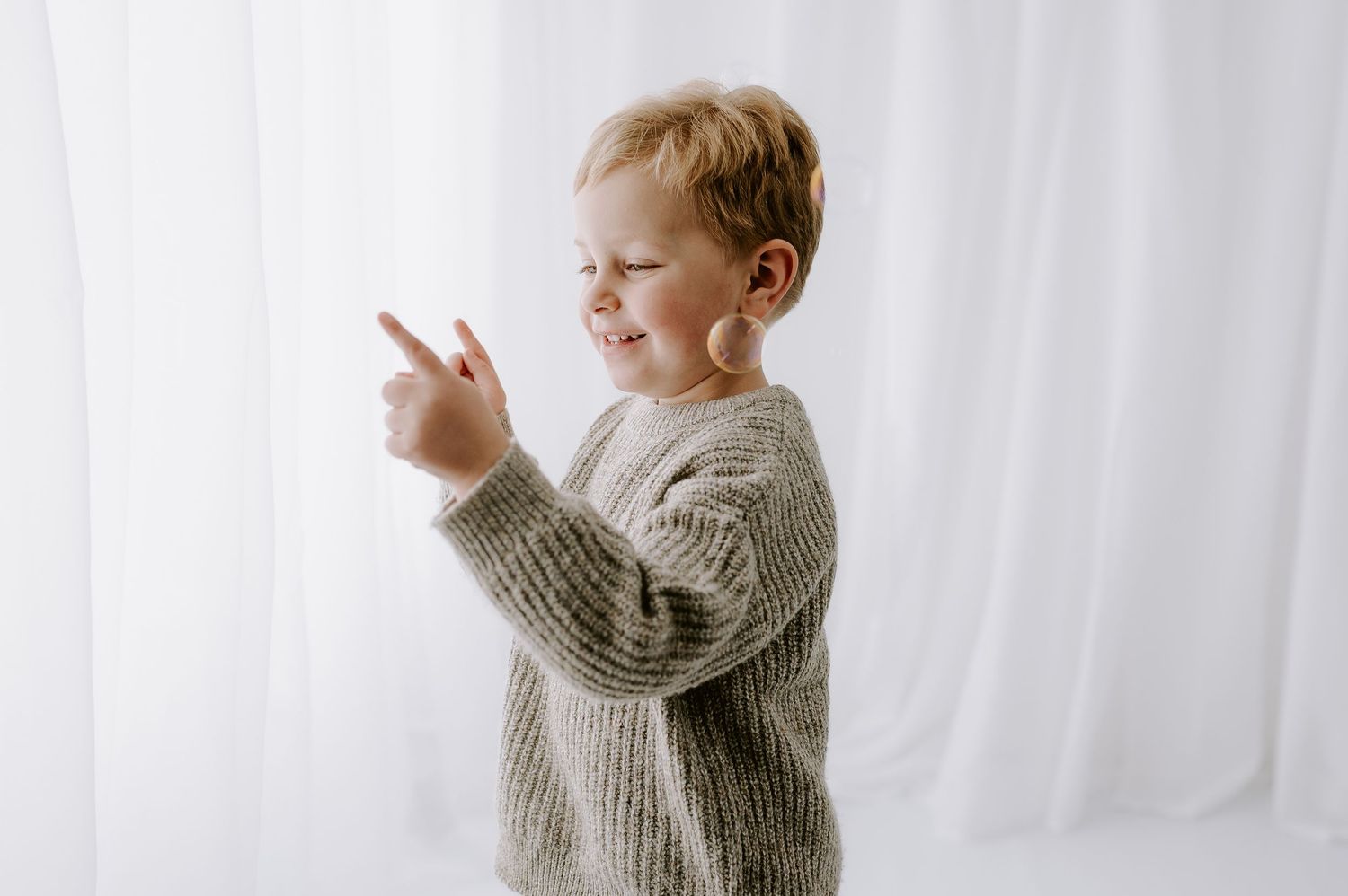 Young child in gray knit sweater playing against white curtain. in bright studio, Melton Mowbray.