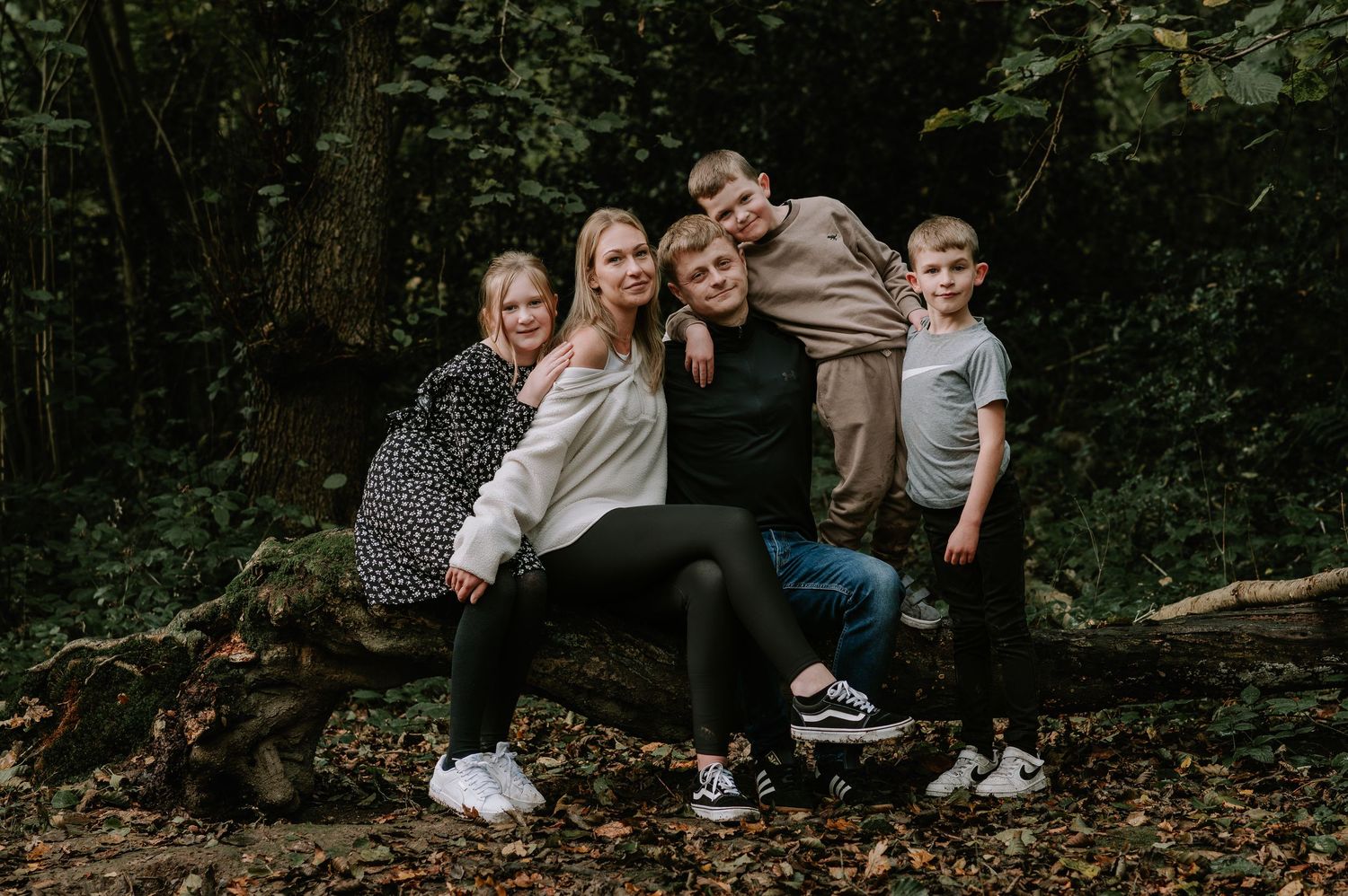 Family of five posing together on a fallen log in a lush green forest setting.