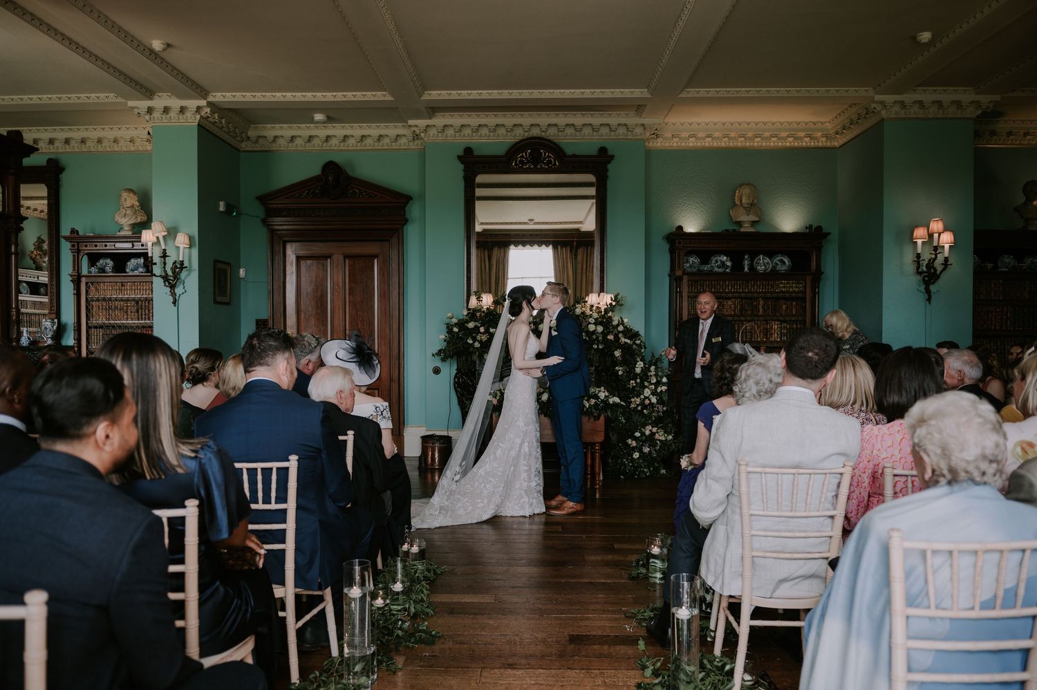 Indoor wedding ceremony in a historic room with turquoise walls, wooden floors, and guests seated in white chairs.