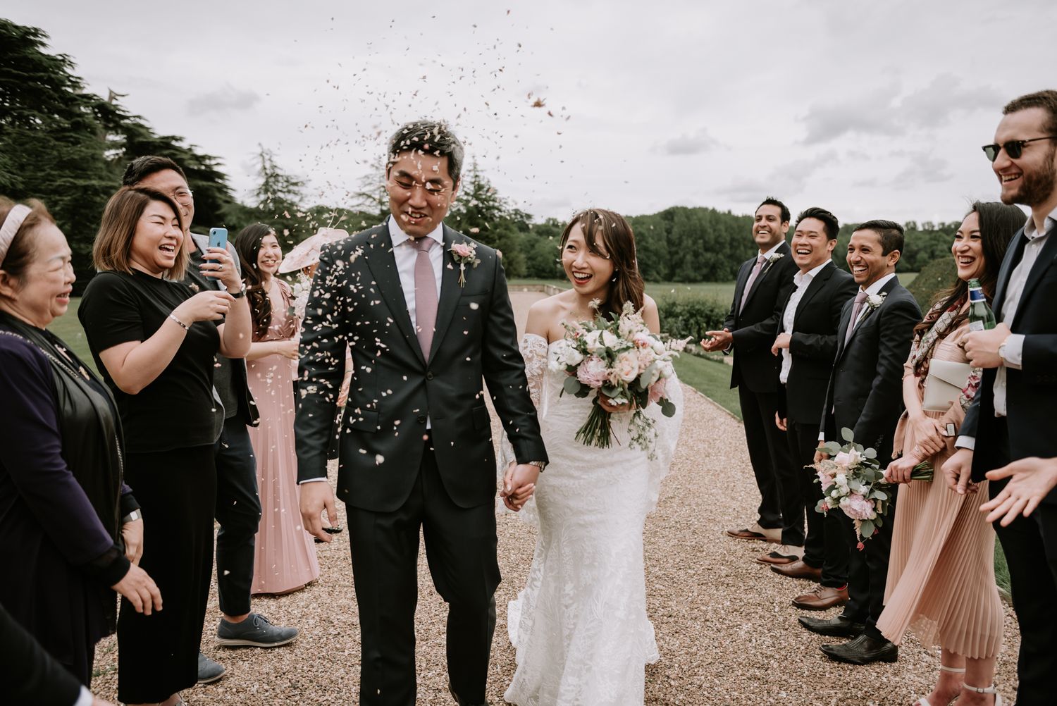 Wedding party guests throw confetti over newlyweds walking down path after ceremony on overcast day.