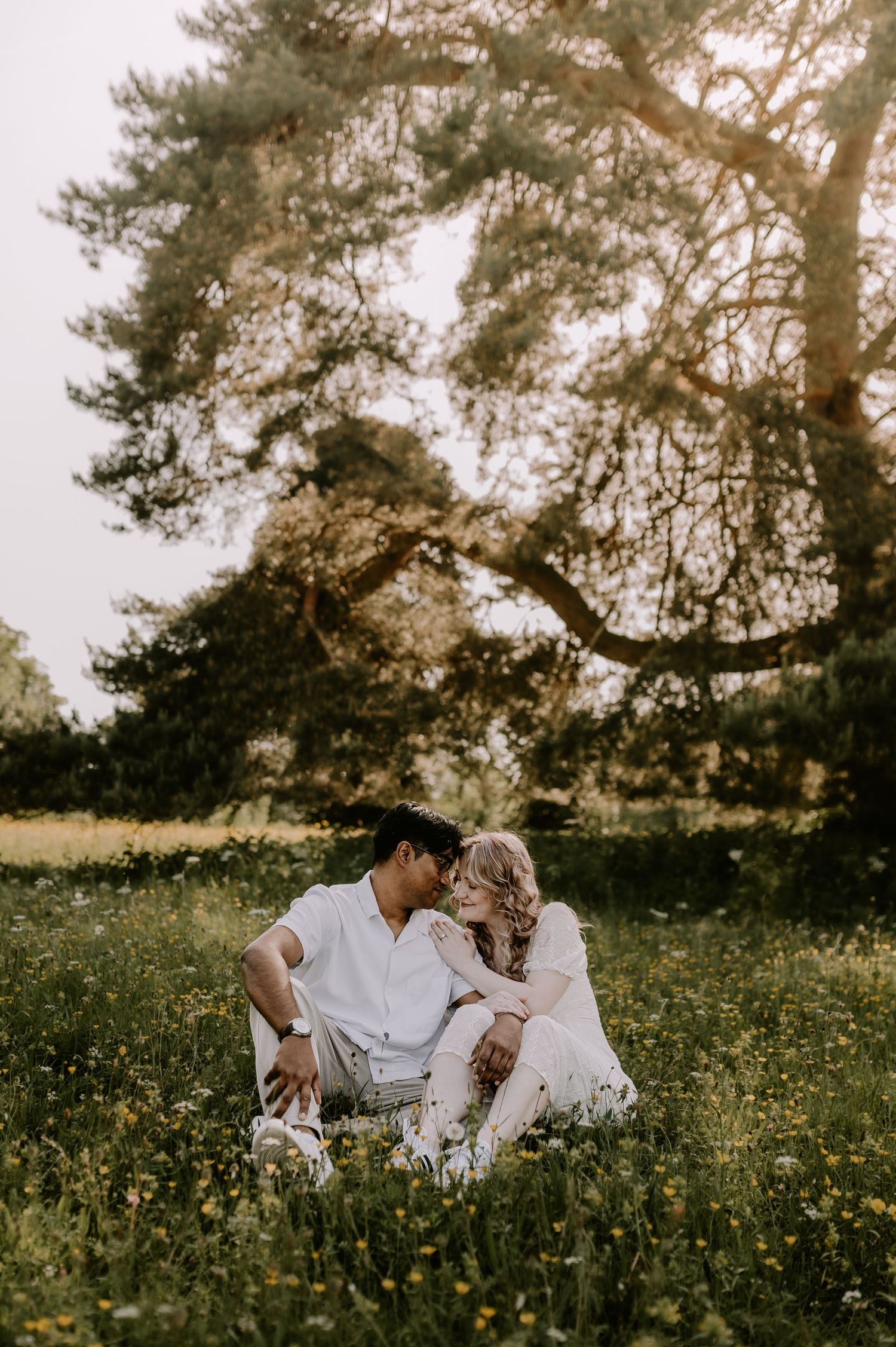 A couple sits romantically in a sunny wildflower meadow, leaning close together beneath a large tree.