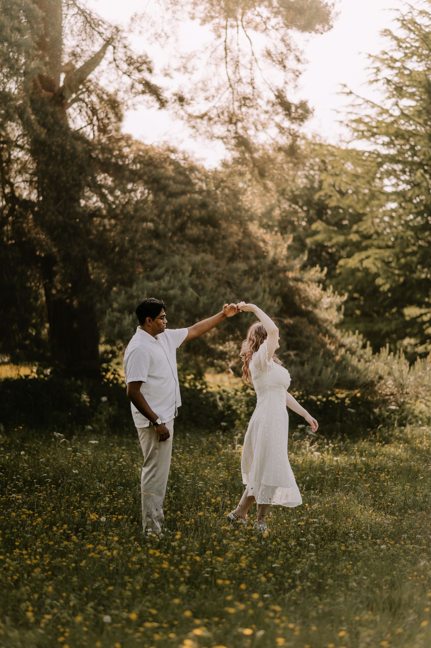A couple dances together in a sunlit meadow with wildflowers, the woman in a white dress being twirled by her partner.