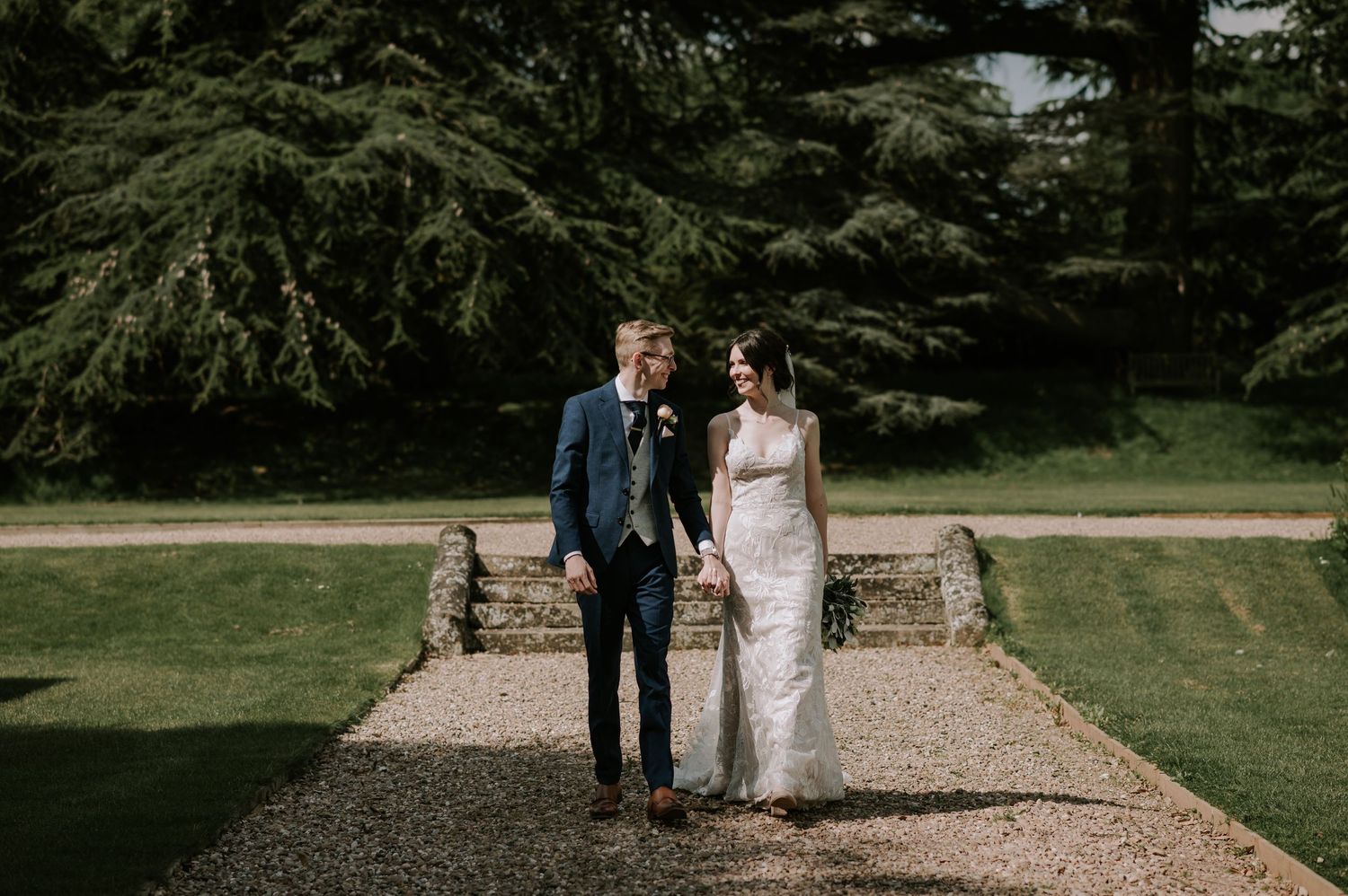 A couple walks hand in hand along a gravel pathway surrounded by lush green gardens on their wedding day.